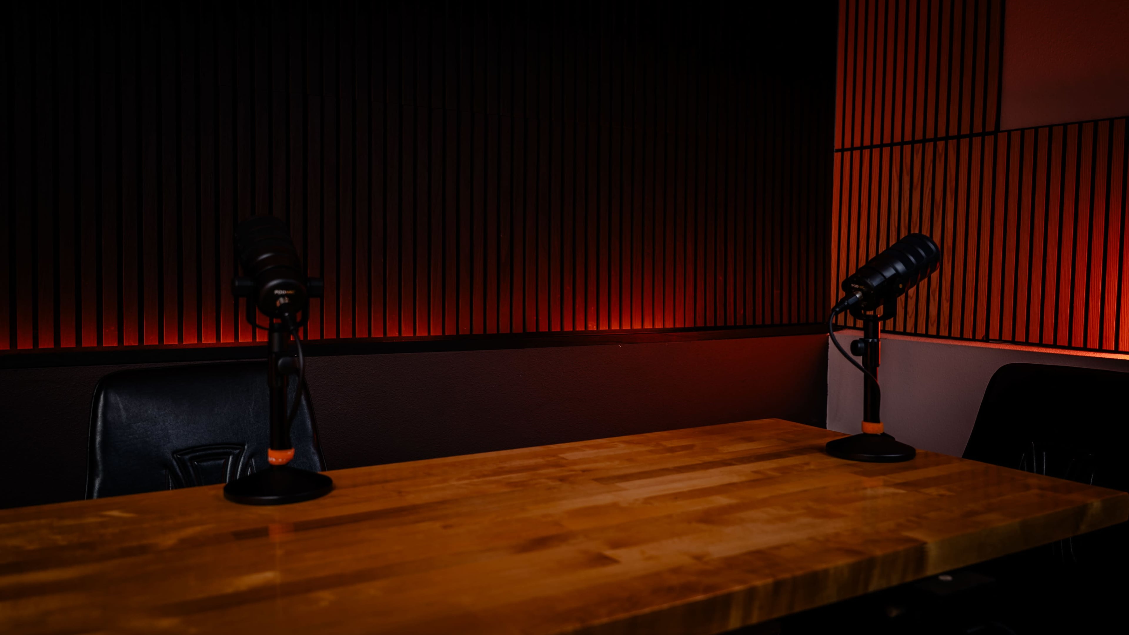 A wooden table with two microphone stands is positioned in a dimly lit recording studio featuring vertical wooden paneling.