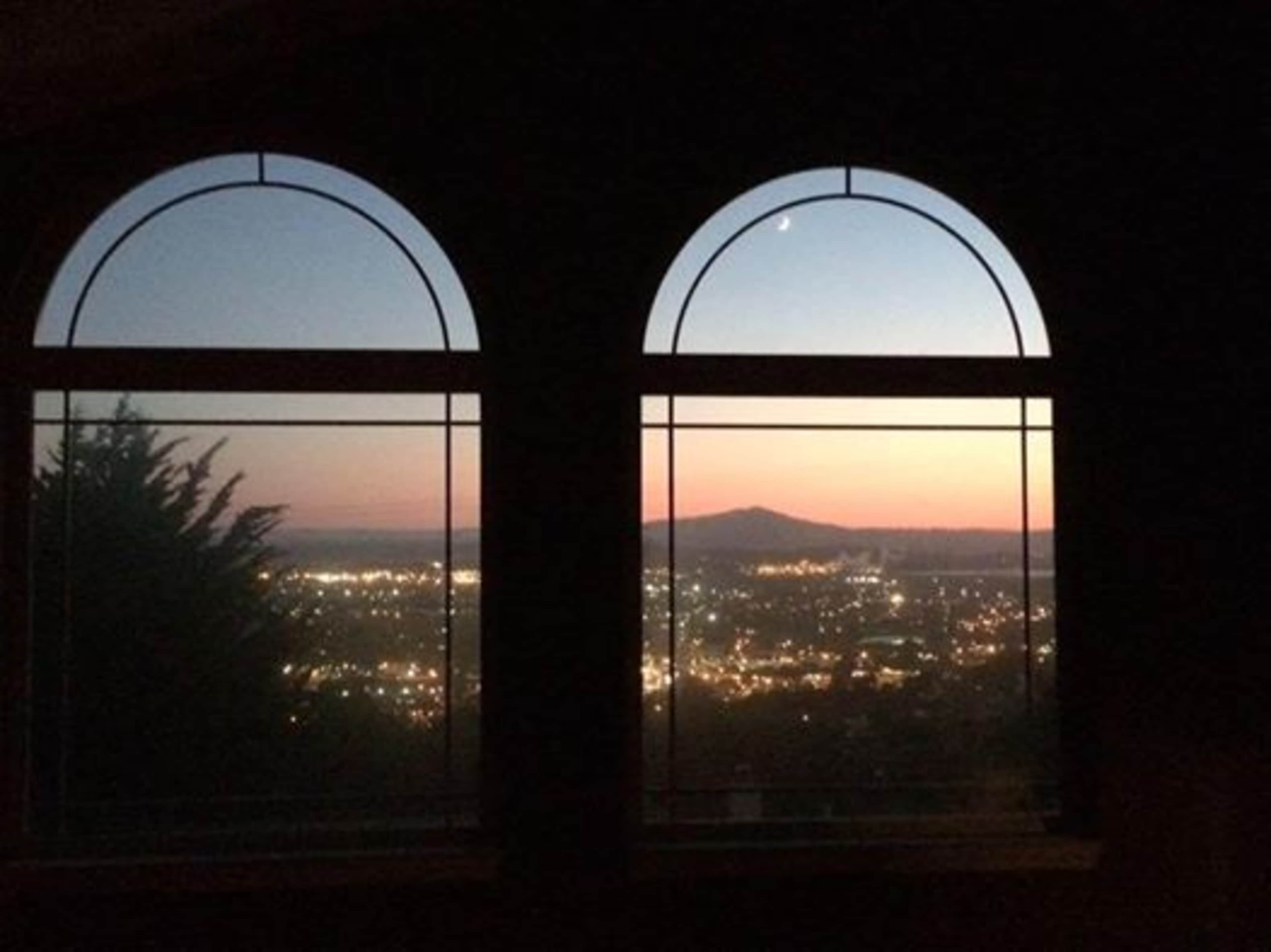 The image shows two arched windows framing a city skyline at dusk, with twinkling lights and a mountain in the background.