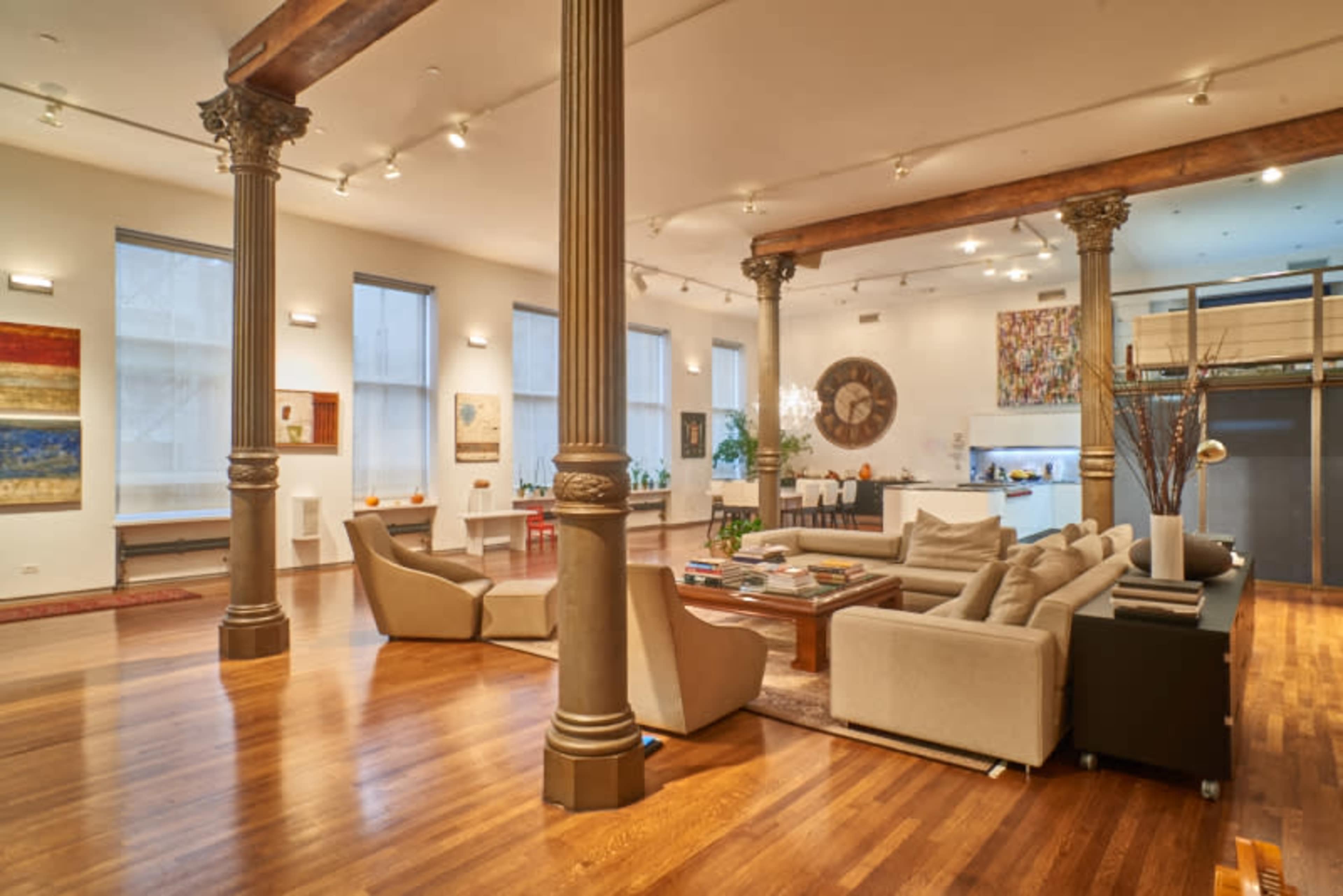A spacious, modern living area with tall columns, wooden floors, and large windows, furnished with a sectional sofa and various artworks on the walls.