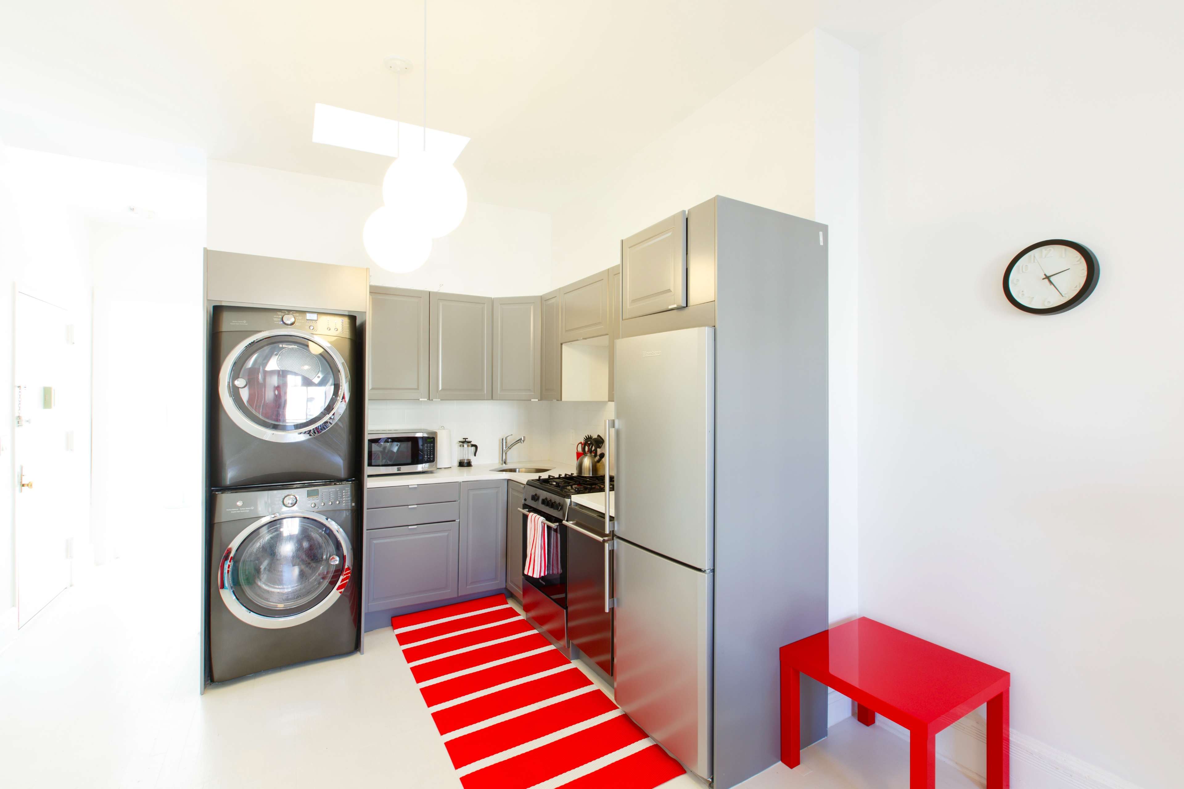 A modern laundry area features stacked gray washers and dryers, a silver refrigerator, a black microwave, and a red rug, all within a bright, minimalist kitchen space.
