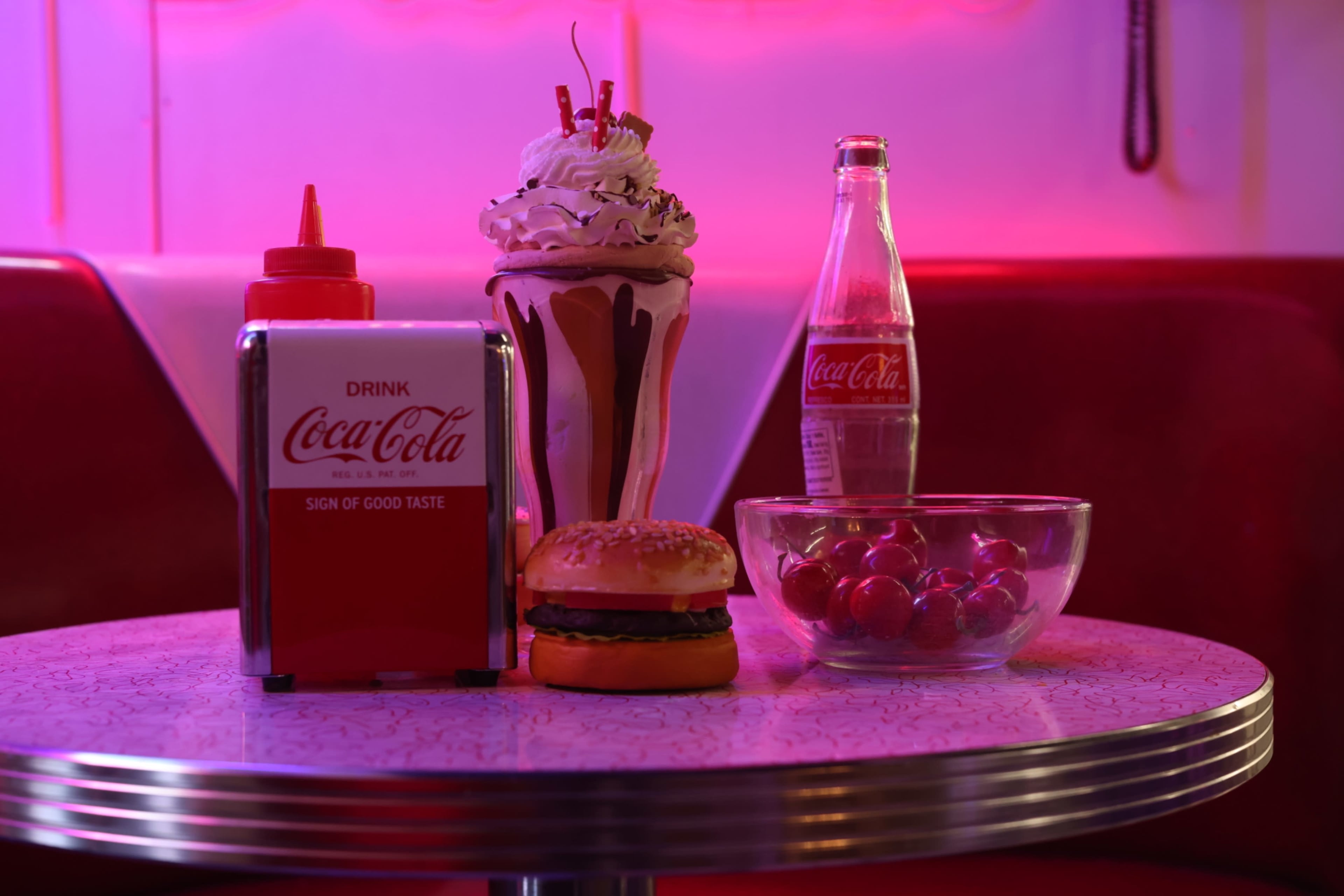The image shows a retro diner table set with a bottle of Coca-Cola, a glass of milkshake topped with whipped cream, a cheeseburger, and a bowl of cherries.