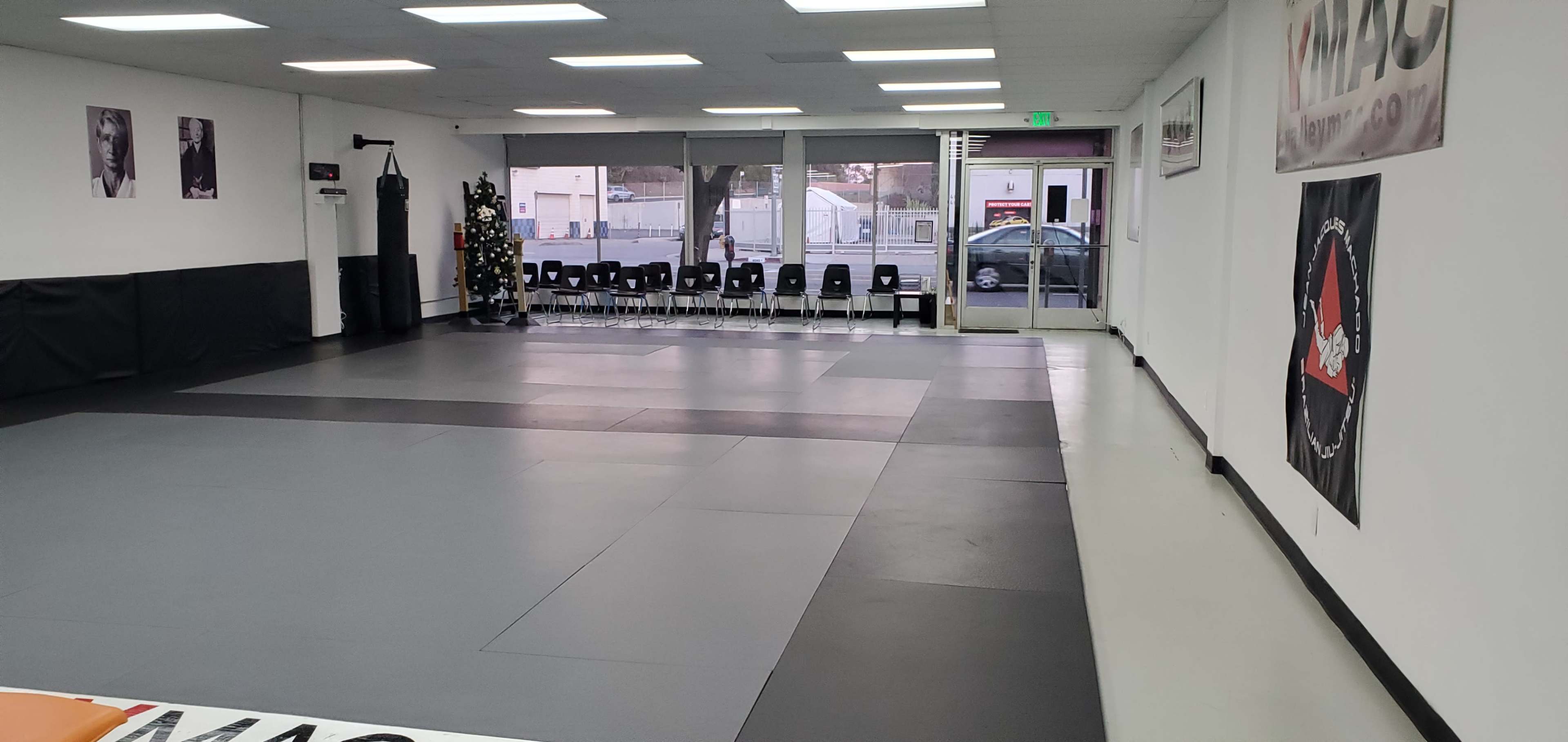 A spacious martial arts studio with gray mats, empty chairs along one wall, and large windows revealing an outside view.