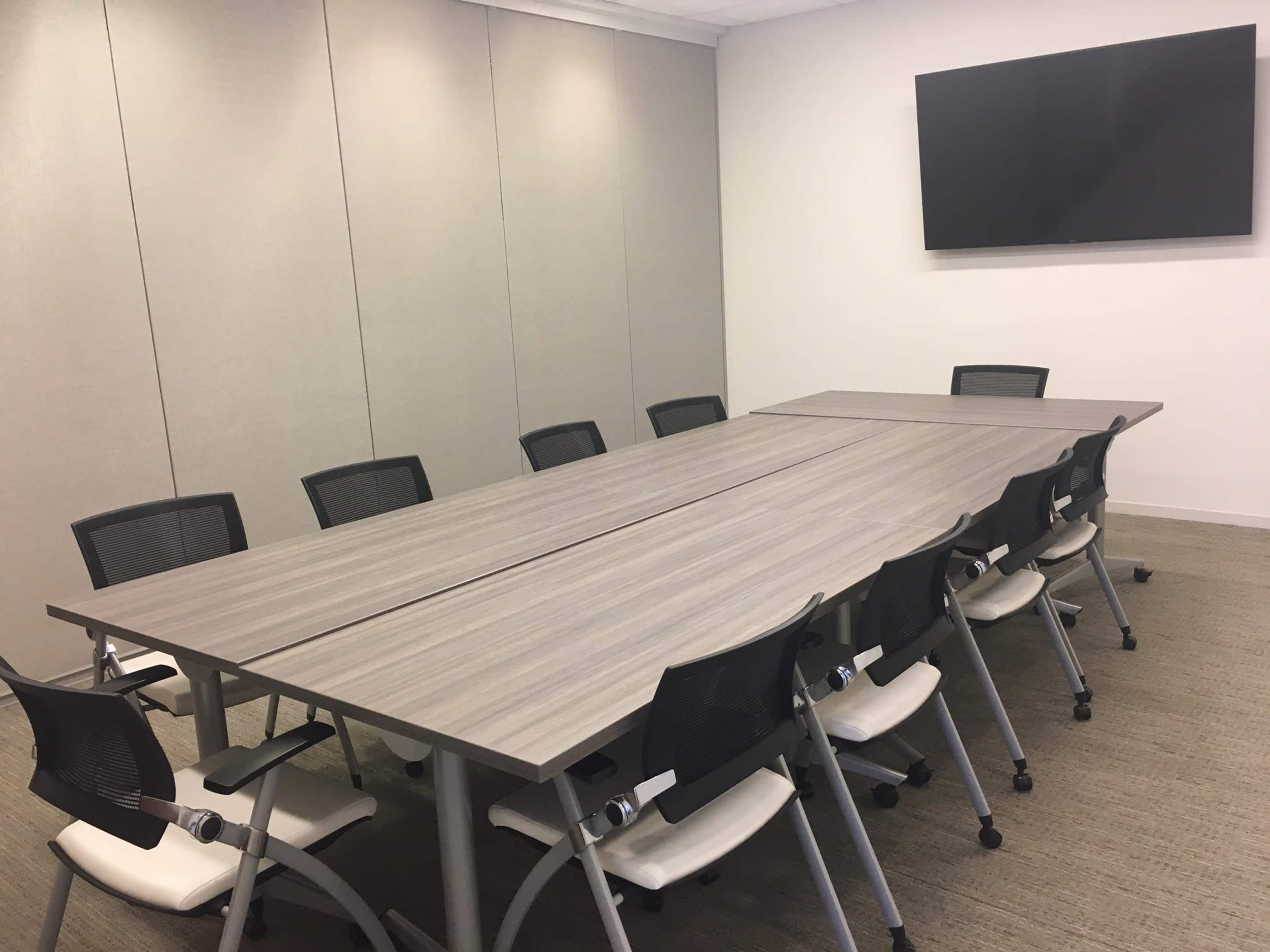 A large conference room features a long rectangular table with ten ergonomic chairs and a wall-mounted flat-screen television.