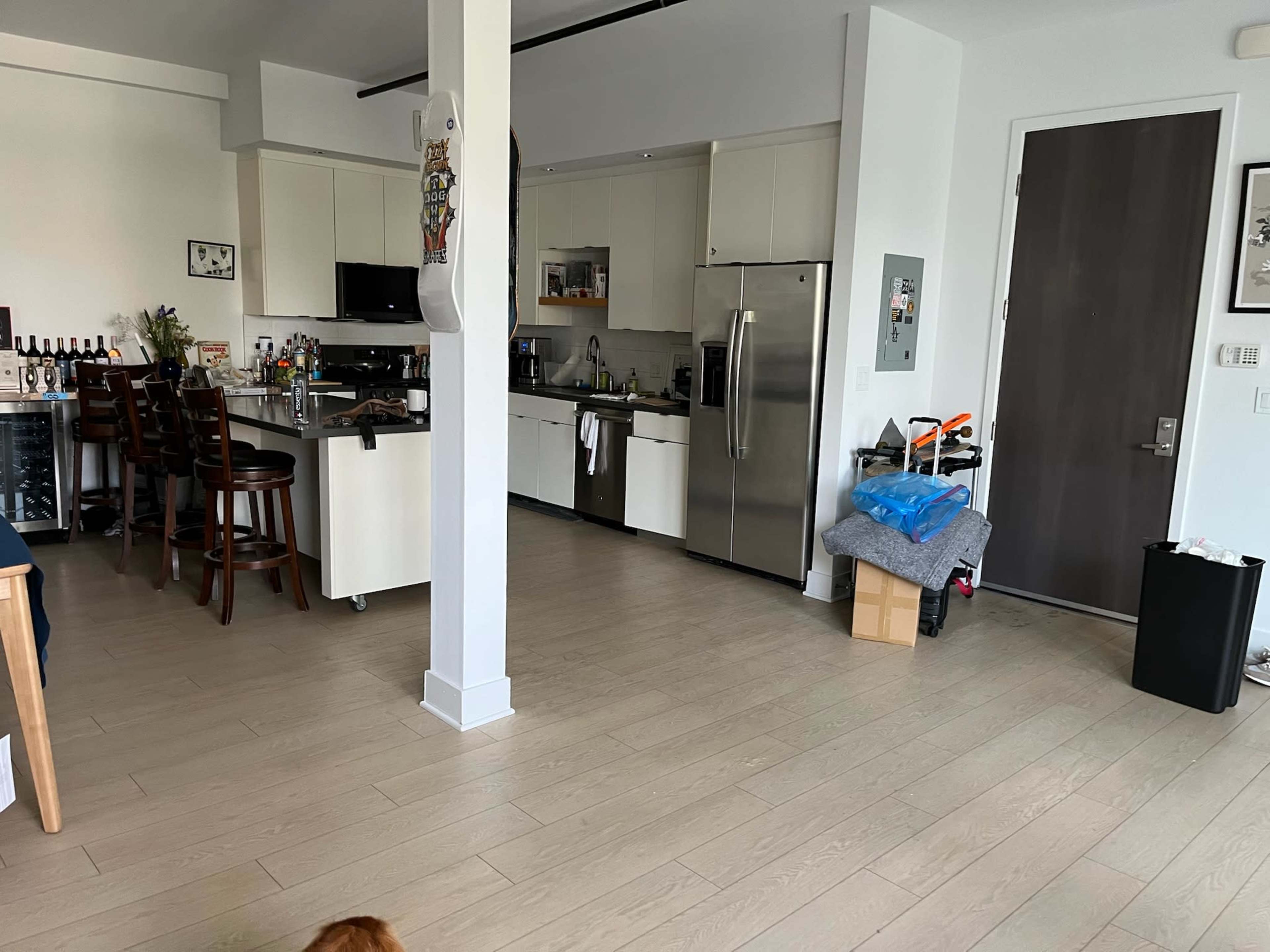 The image shows a modern kitchen and dining area with wooden floors, a stainless steel refrigerator, and various items scattered around.