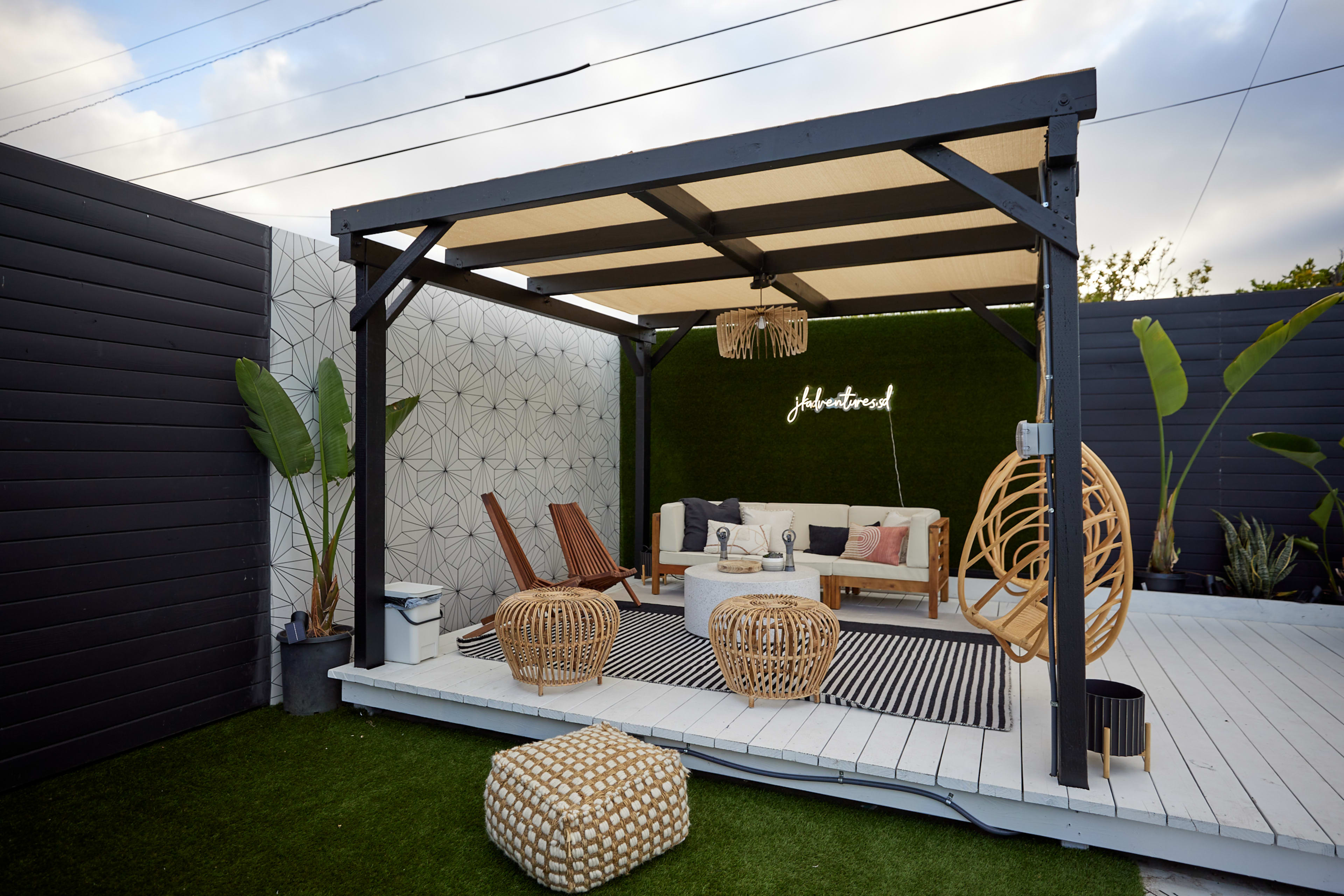 The image shows a modern outdoor patio with a wooden pergola, styled seating area, and decorative plants.