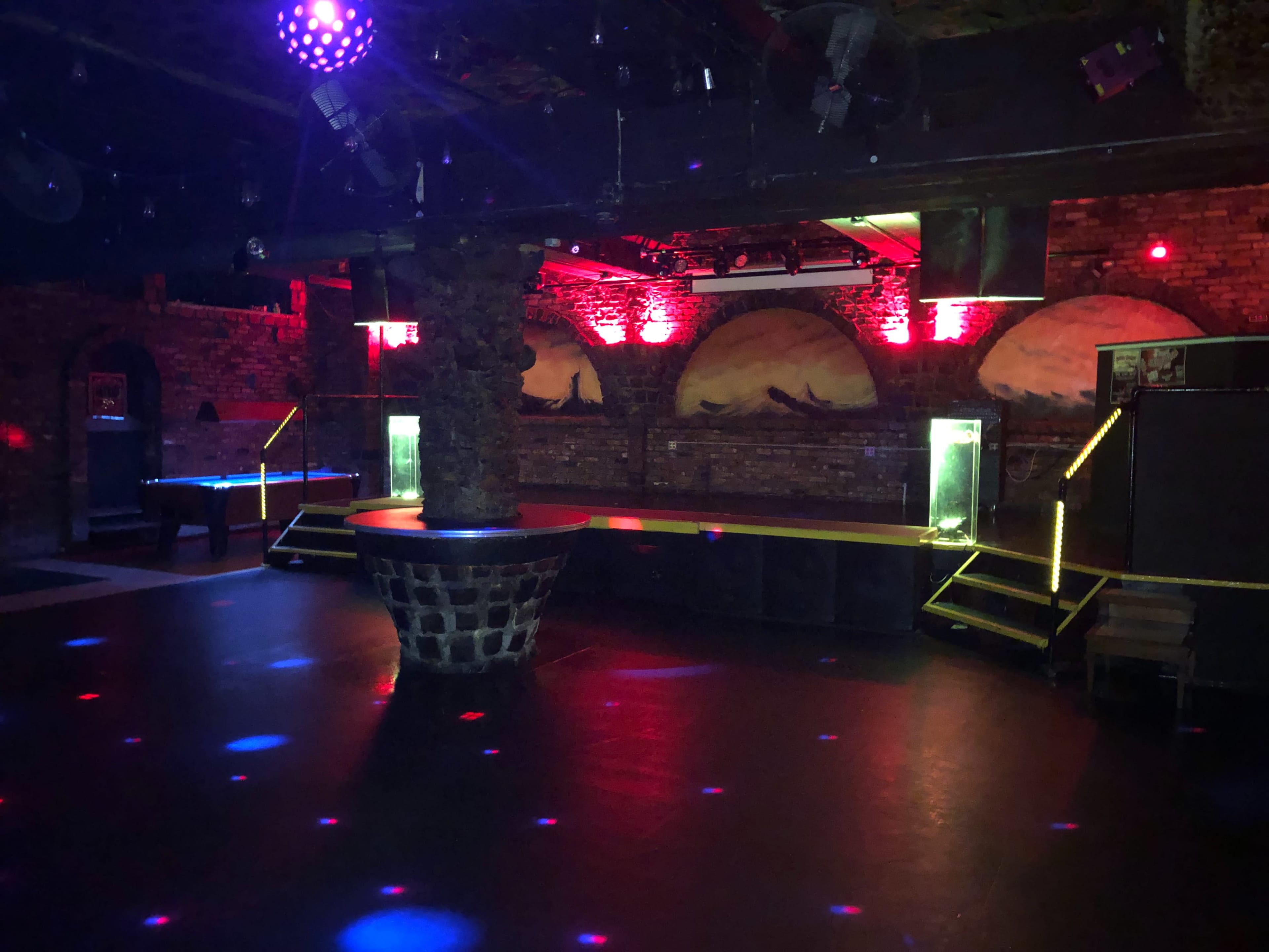 A dimly lit nightclub interior with a circular bar in the center, surrounded by illuminated stages and stone walls.