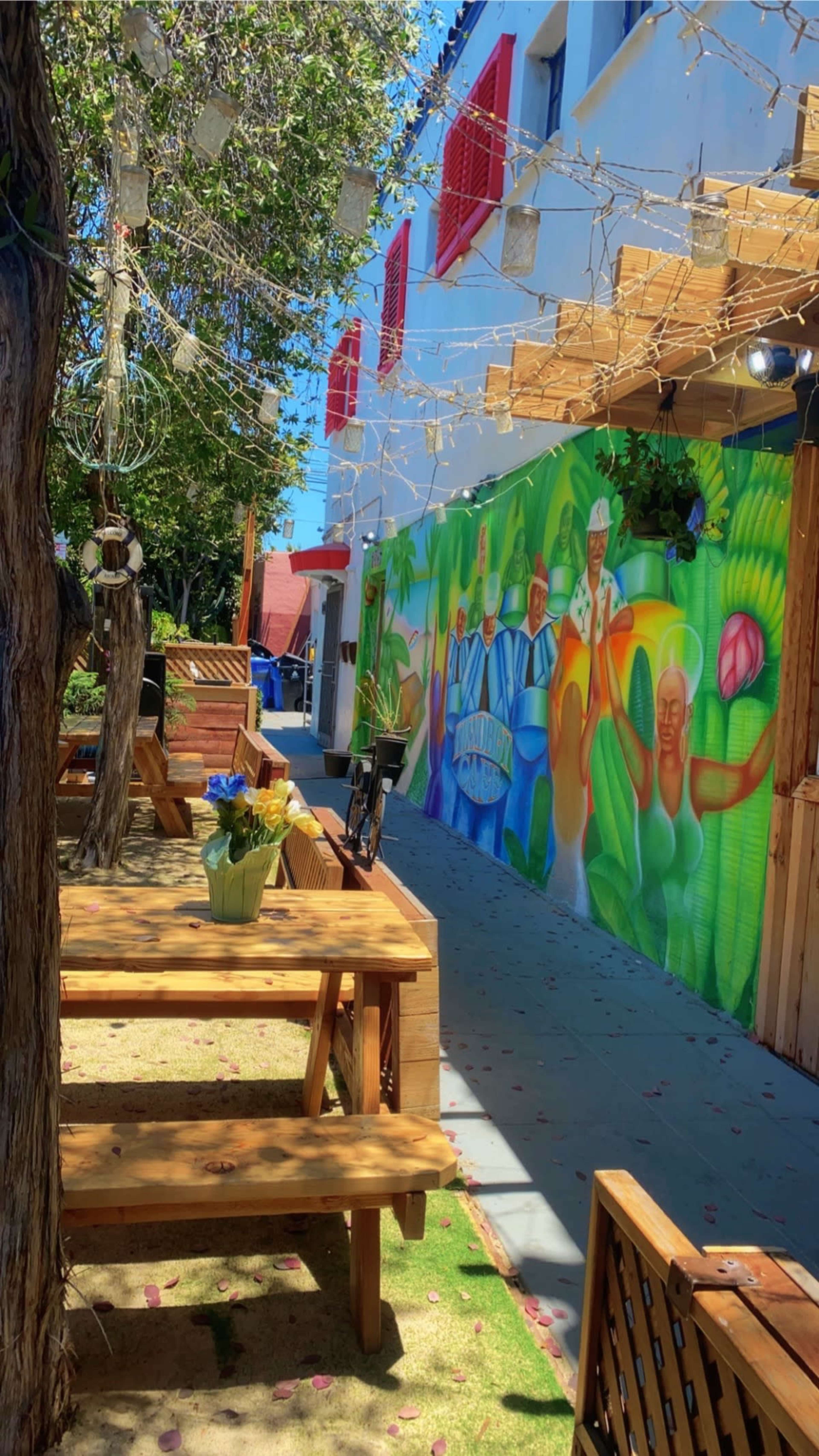 A vibrant outdoor seating area with wooden tables, a mural on the wall depicting colorful figures, and string lights overhead near a landscaped pathway.