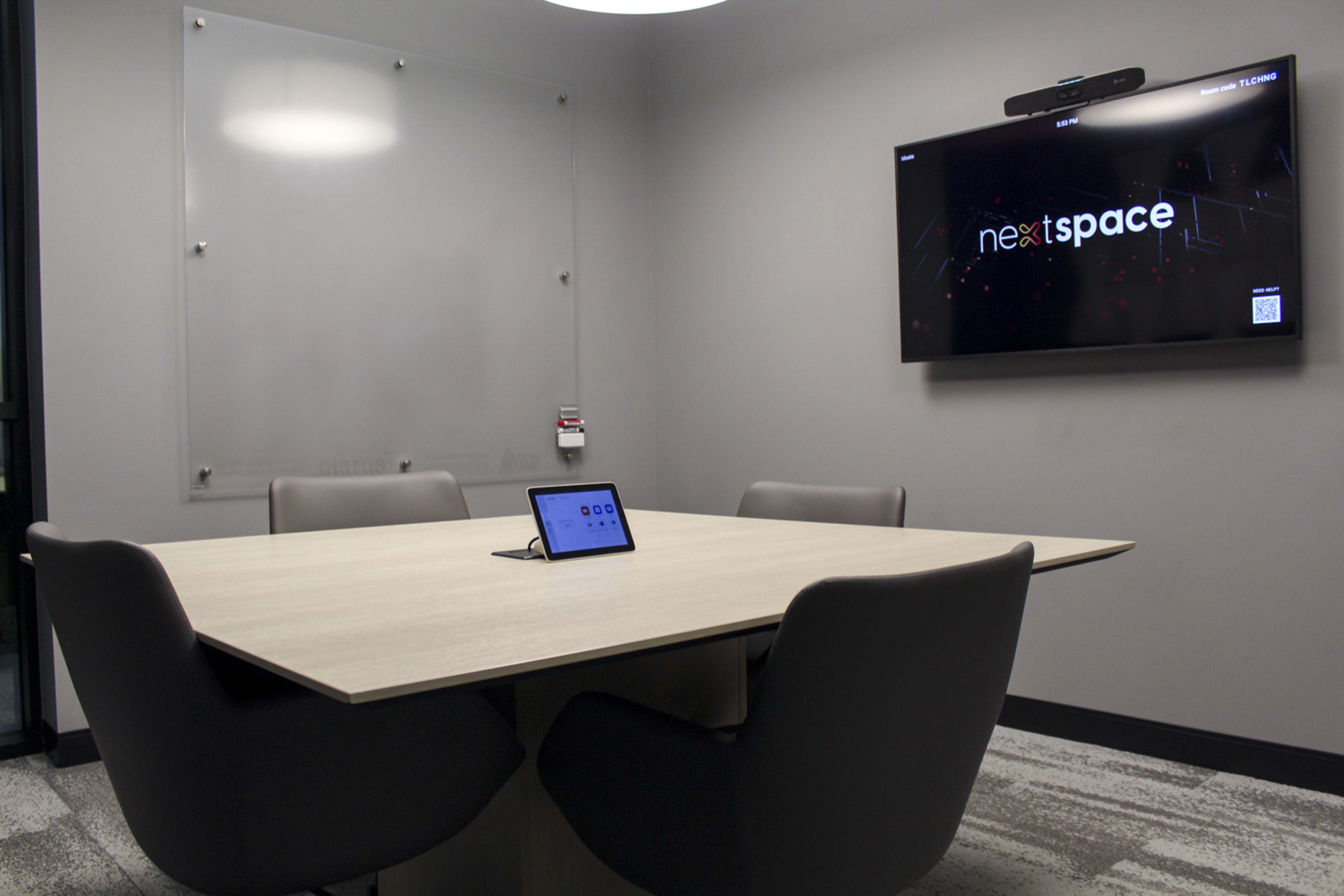 The image shows a modern meeting room featuring a large table, several gray chairs, a wall-mounted screen displaying "nextspace," and a tablet on the table for presentations or video calls.