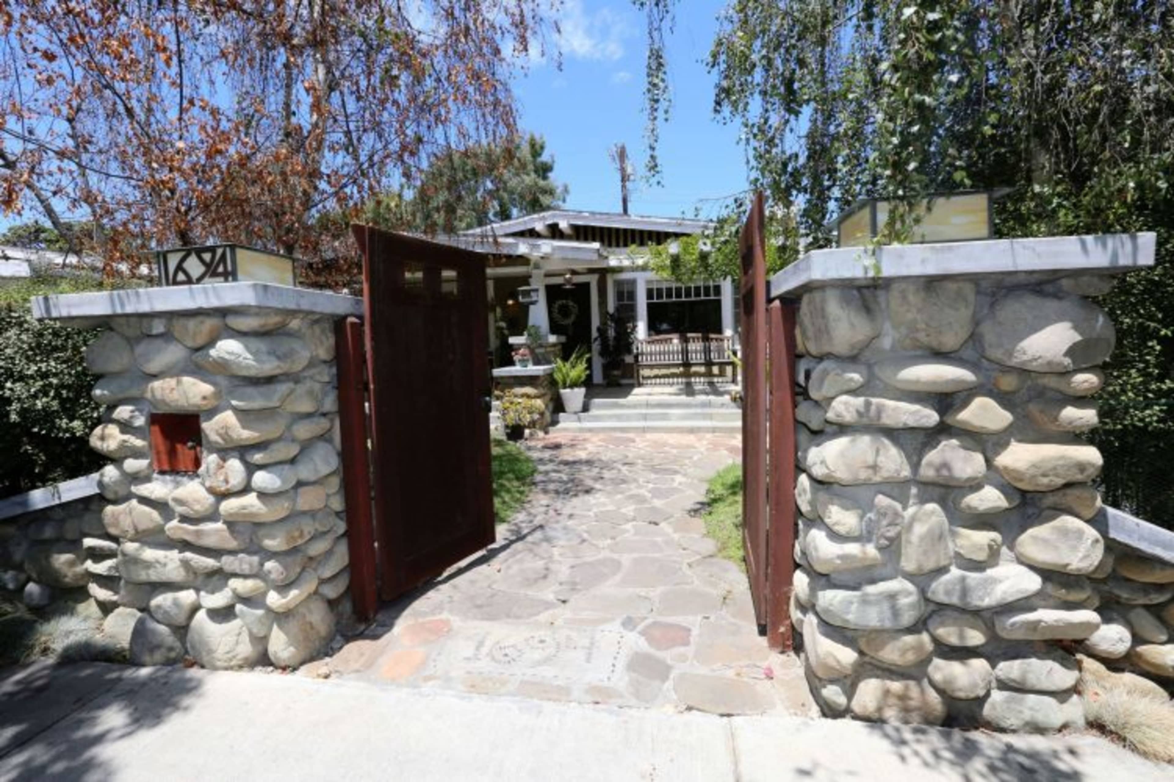 A stone wall with wooden gates opens to a pathway leading to a house surrounded by greenery.