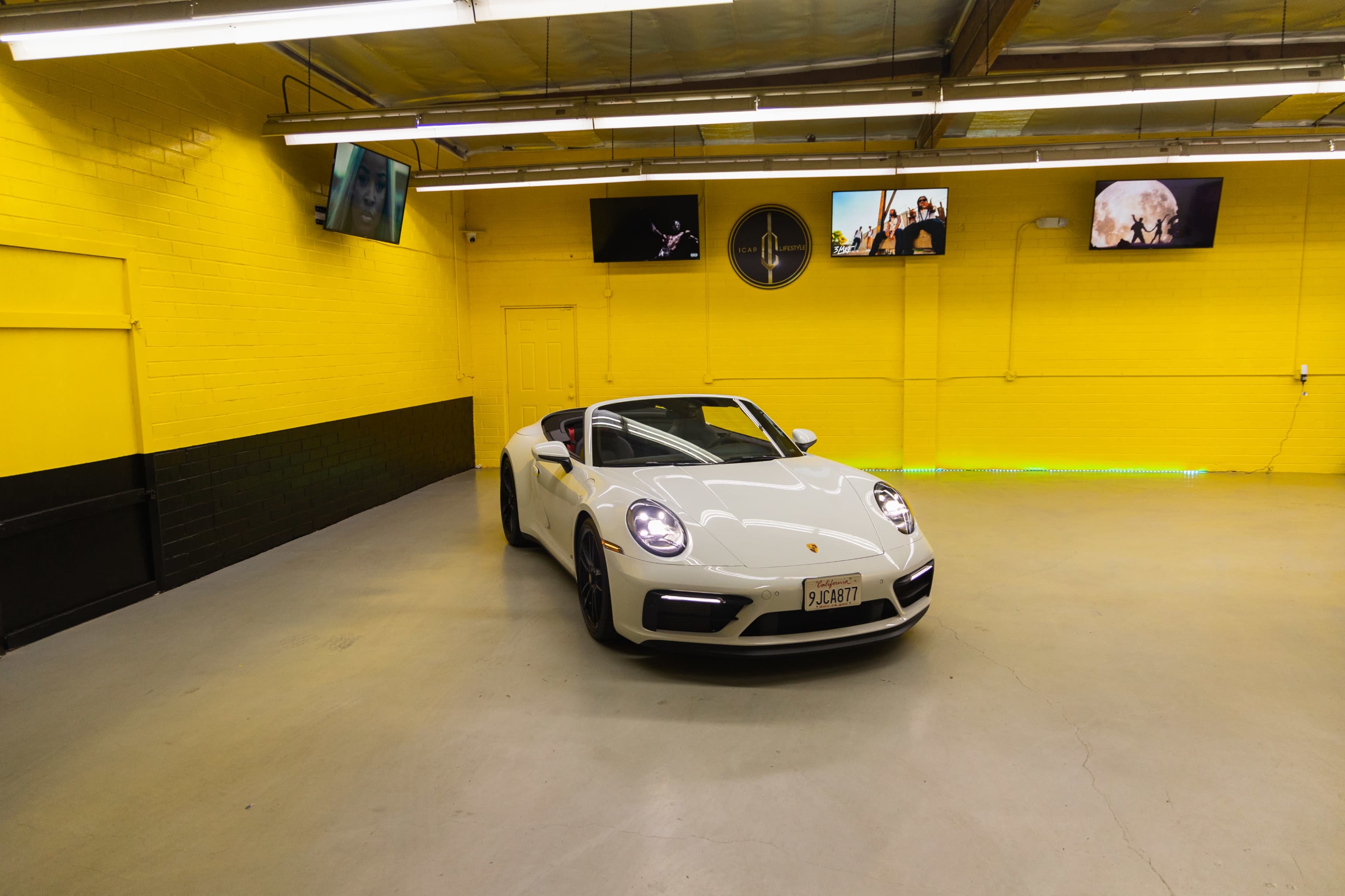 A white Porsche 911 is parked in a yellow-walled garage with multiple screens displaying images.