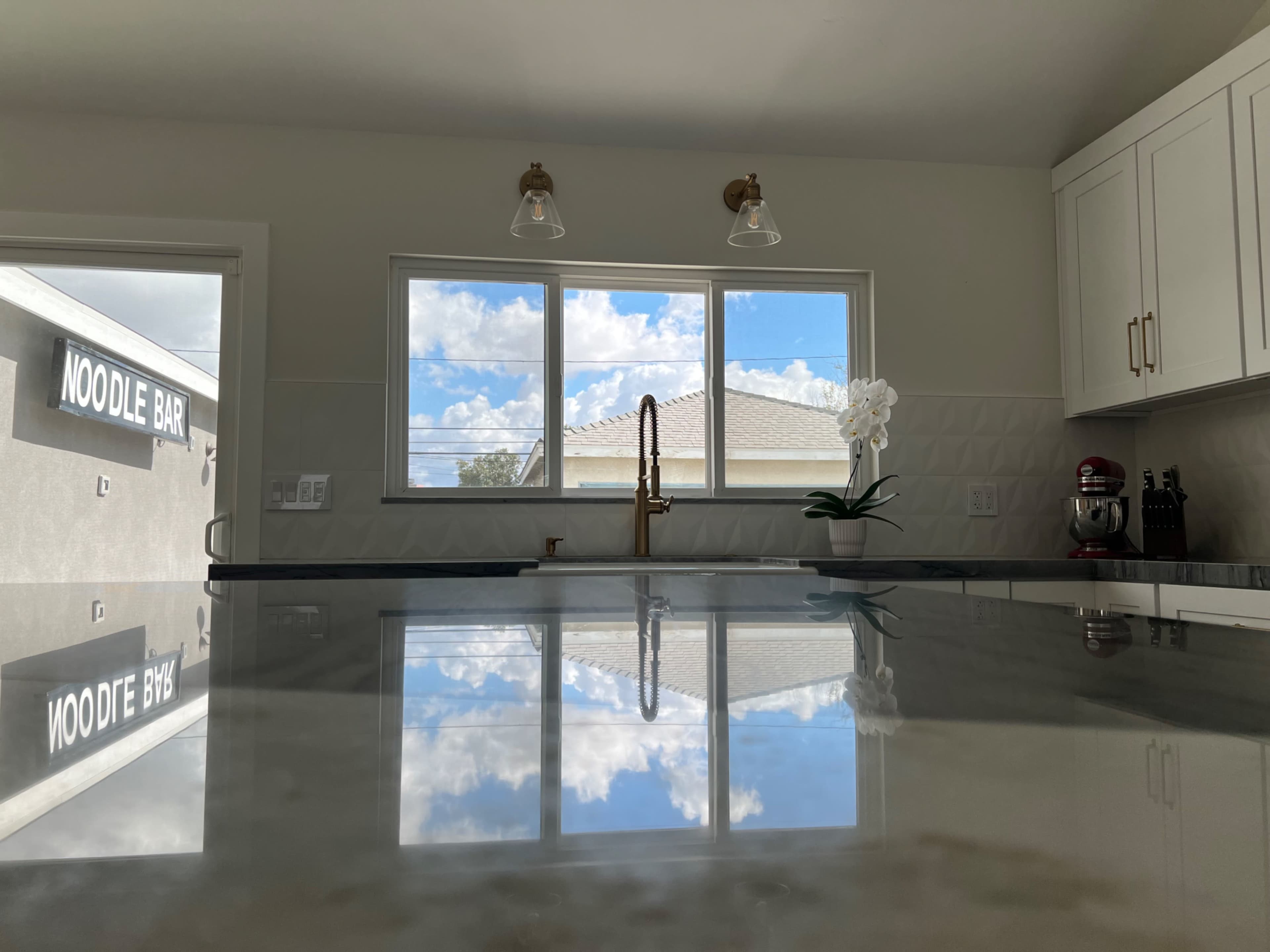 A kitchen with a reflective countertop and large windows showing a blue sky and clouds, overlooking a building labeled "NOODLE BAR."