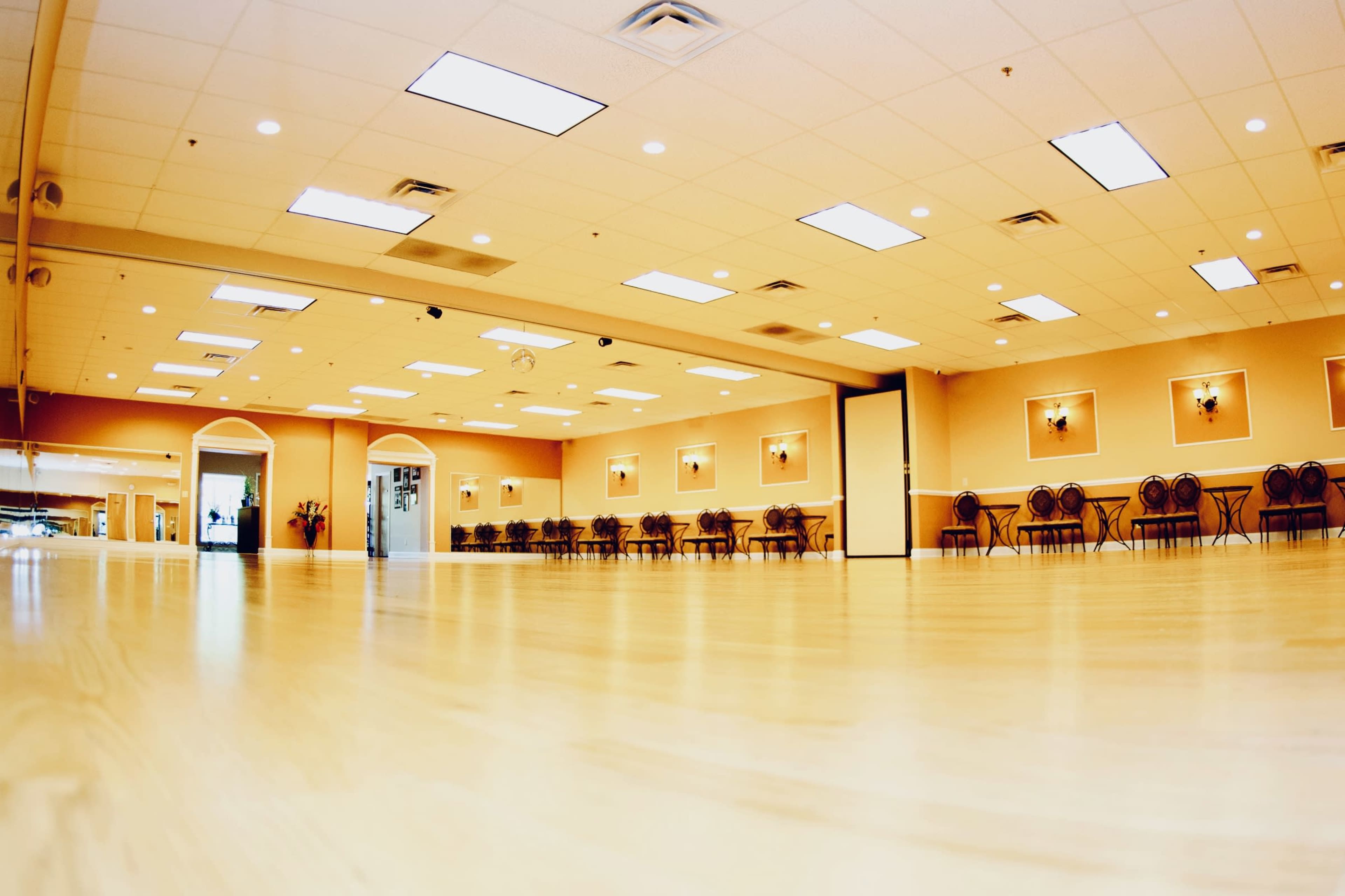 A spacious hall with wooden flooring, illuminated by overhead lights, and lined with chairs along the walls.