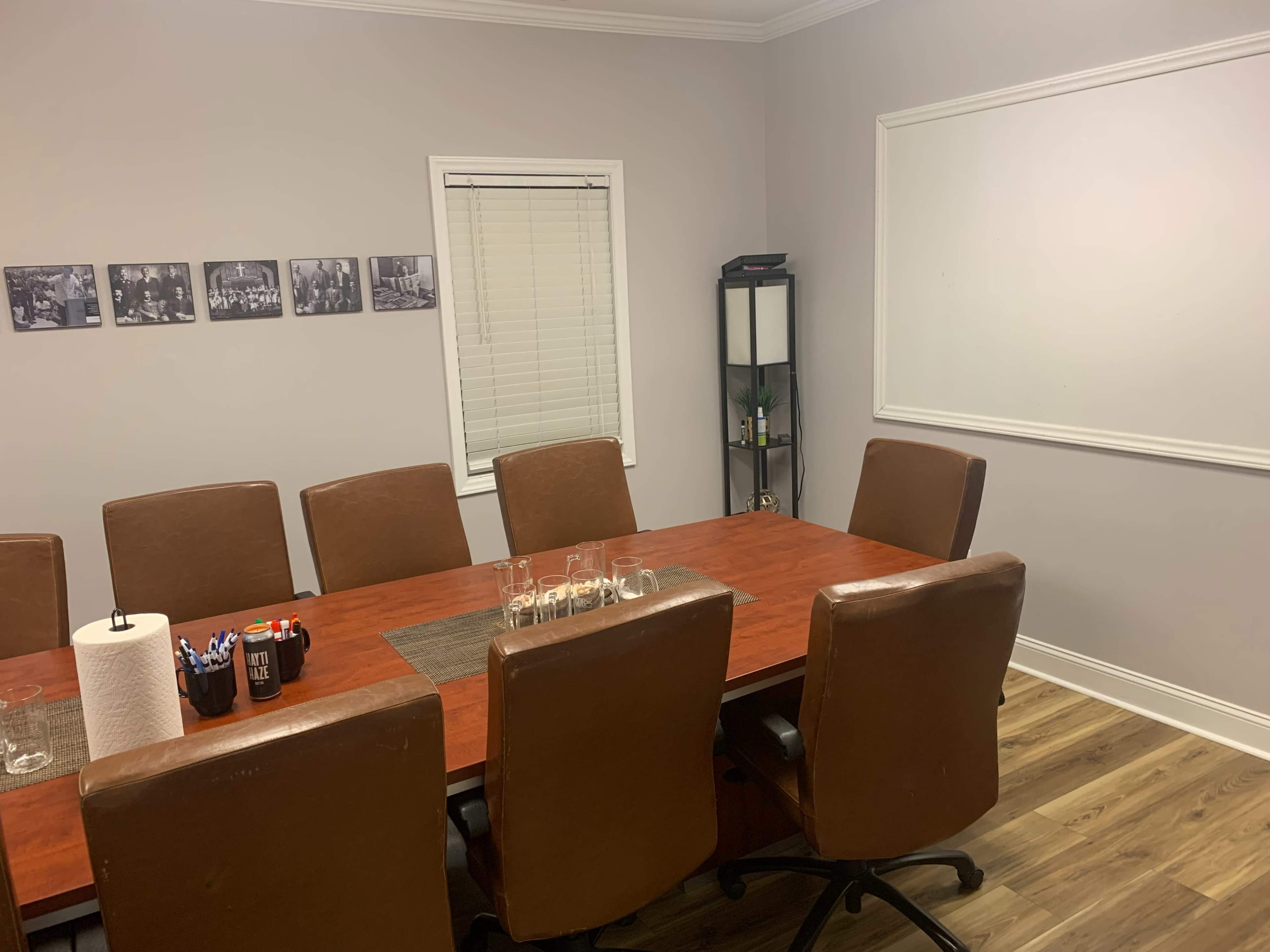 The image shows a conference room with a large wooden table surrounded by eight brown leather chairs, a whiteboard on the wall, and a shelf holding decorative items.