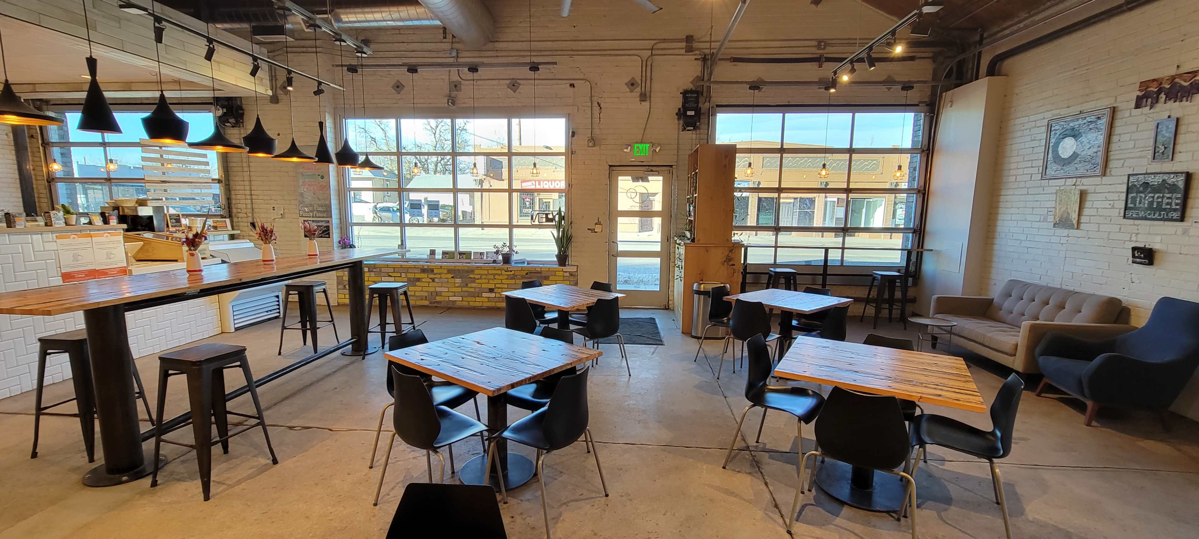 A spacious café with wooden tables, black chairs, a long bar, and large windows offering a view of the street outside.