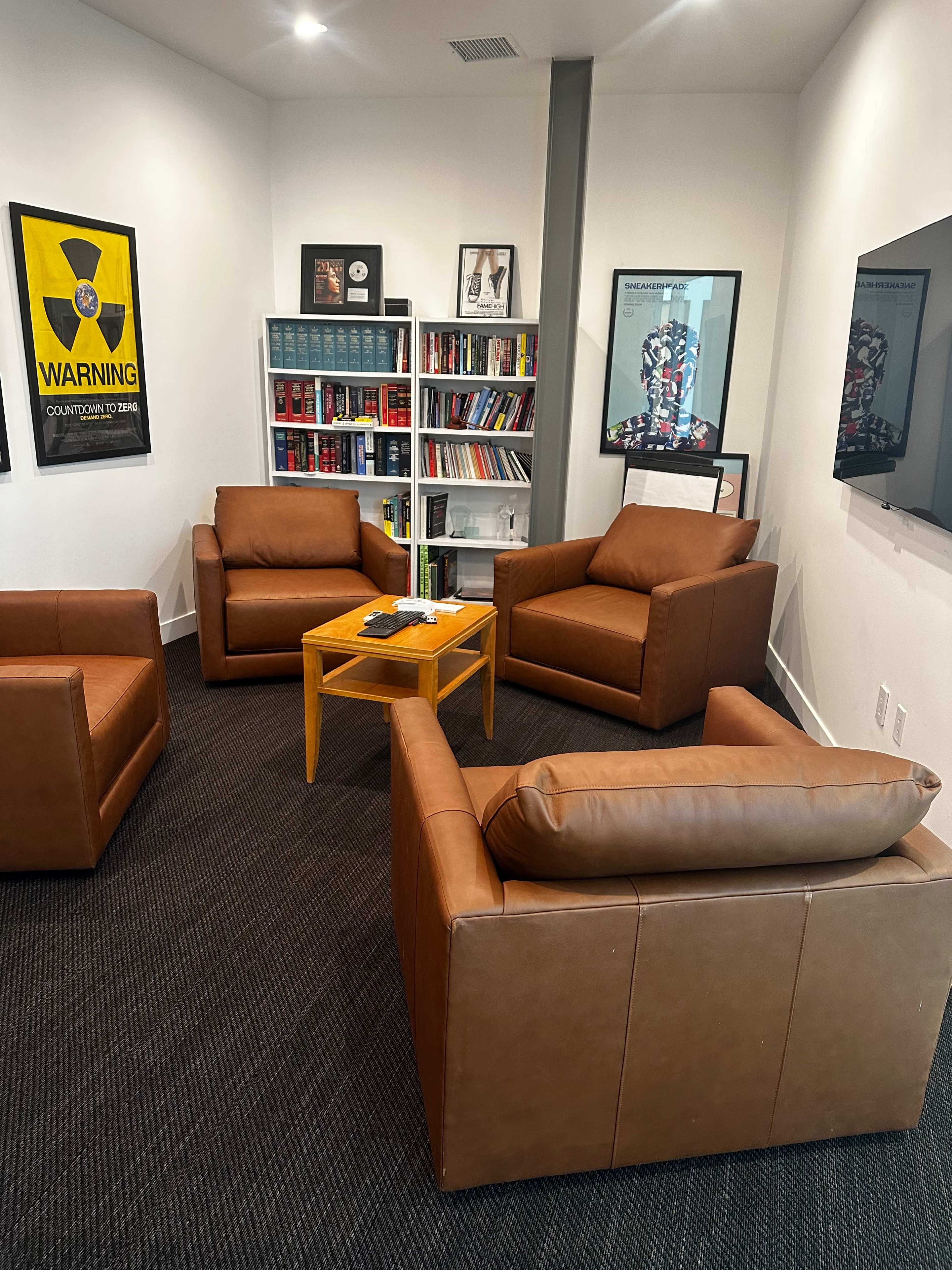 The image shows a modern room with a set of four brown leather chairs arranged around a small wooden table, alongside a bookshelf filled with colorful books and framed artwork on the walls.