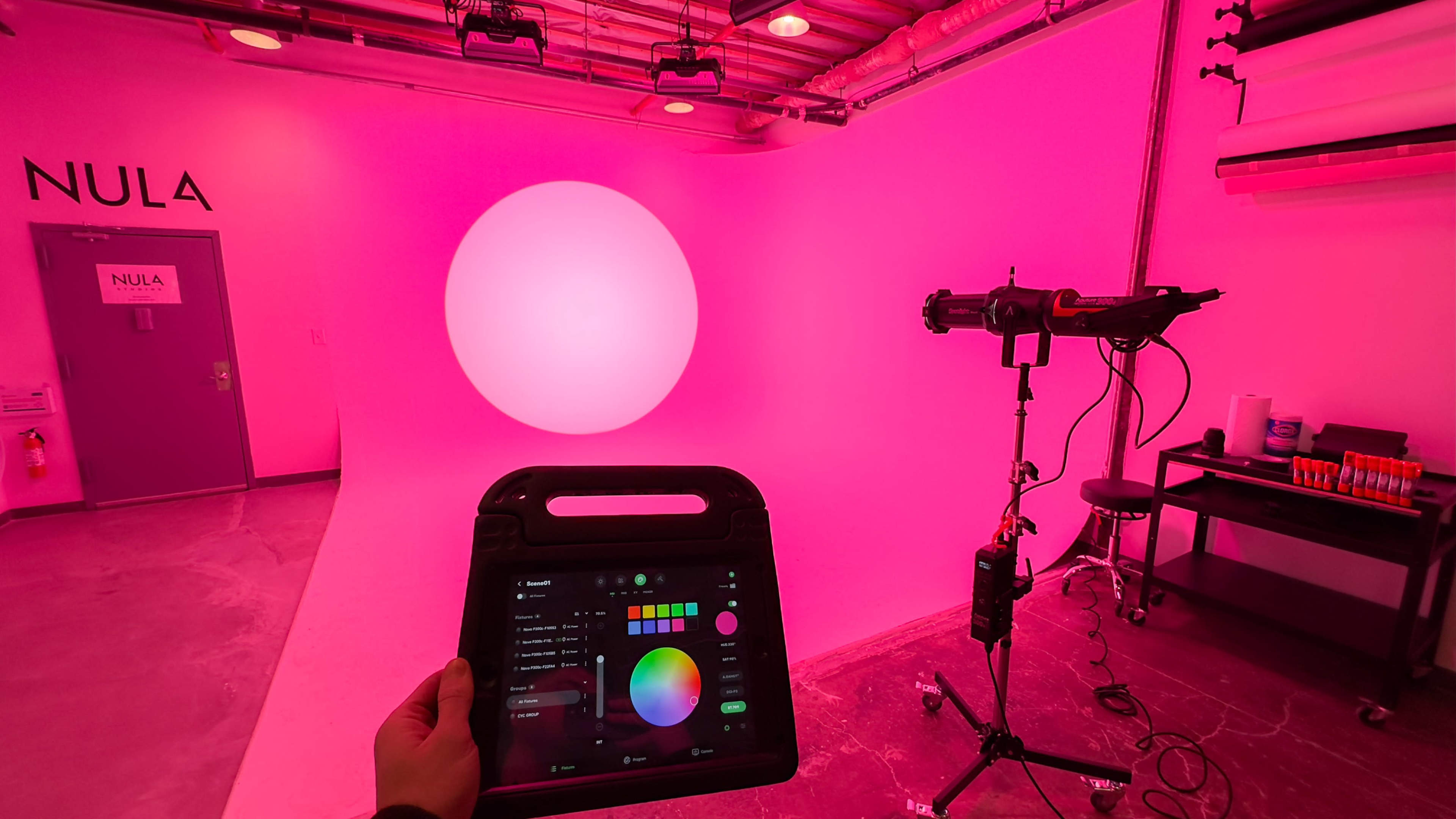 A pink-walled studio features a large illuminated circular light, with a control panel being held in front.