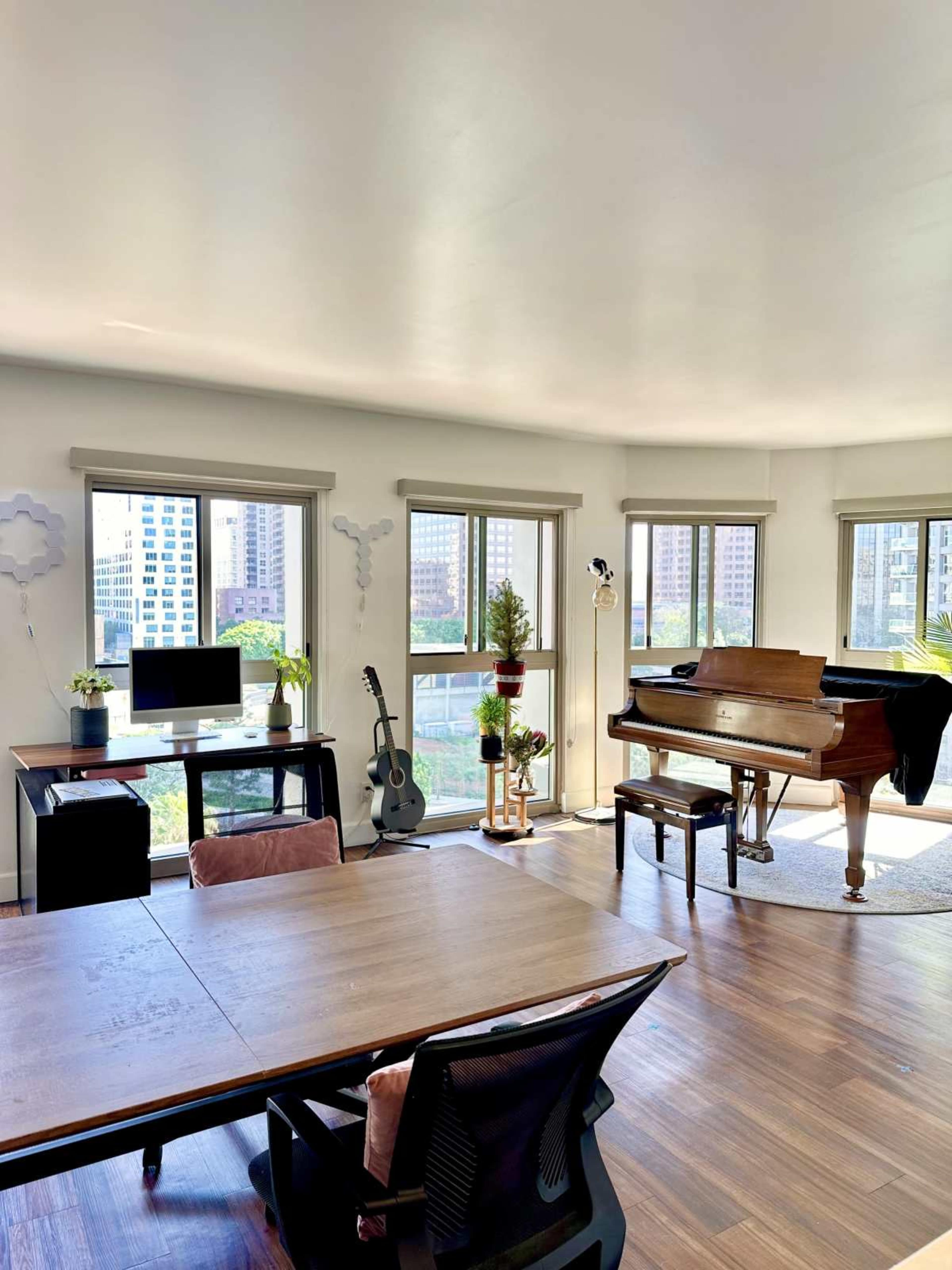 The image shows a bright, modern room featuring a piano, a desk with a computer, an acoustic guitar, and multiple large windows allowing natural light to flood the space.