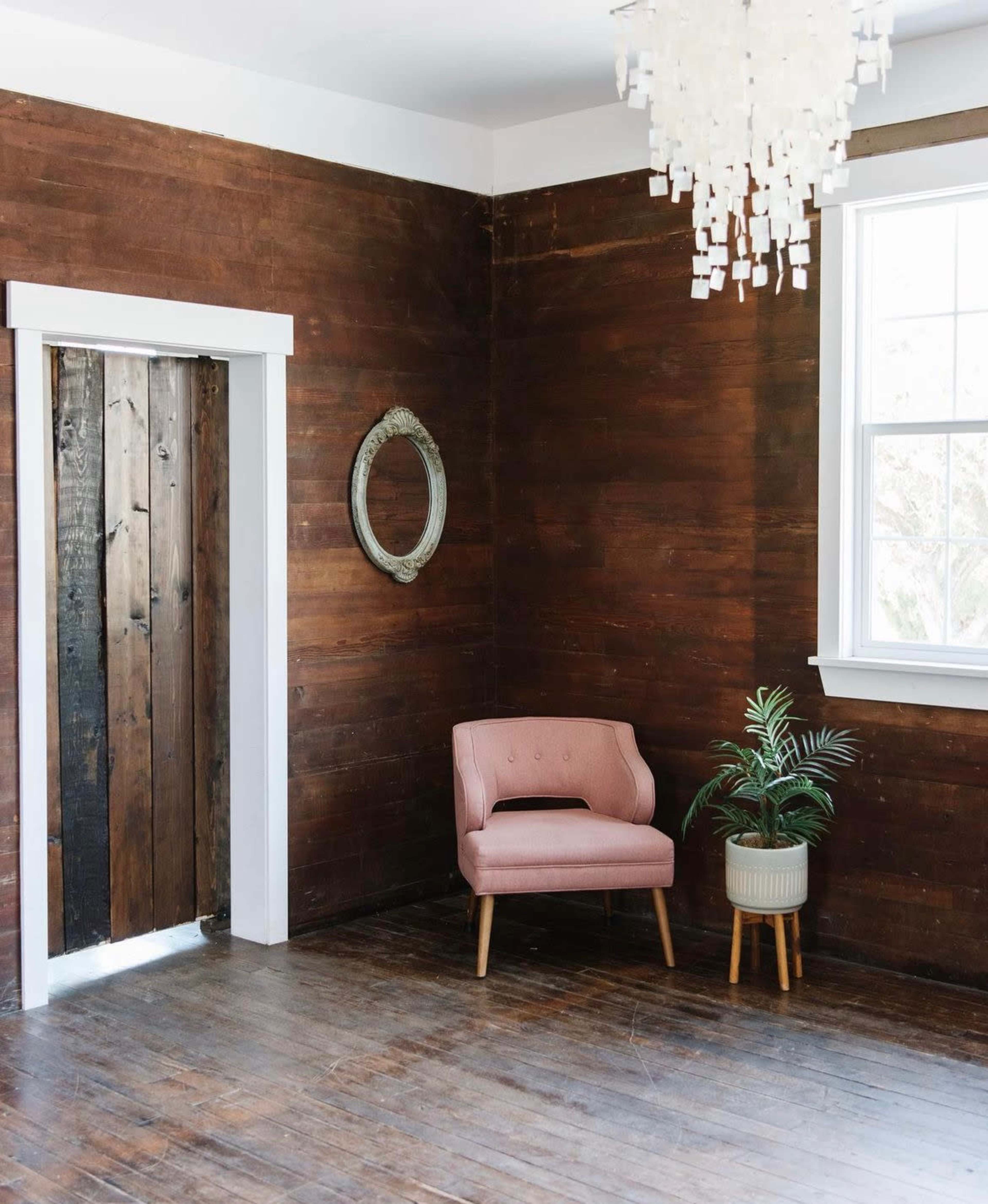 A pink chair is positioned next to a window in a room with wooden walls and a wooden door.