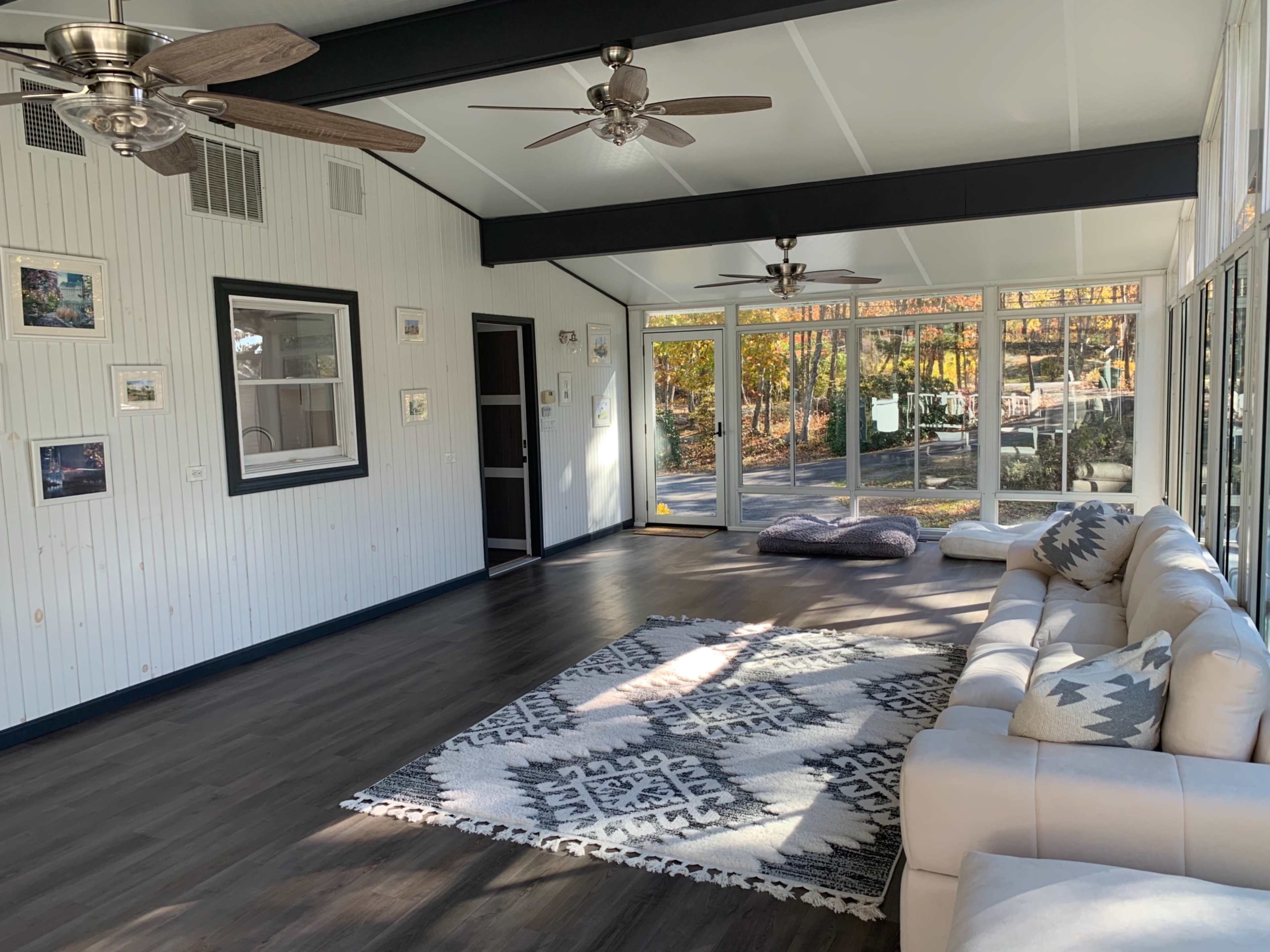 A bright sunroom with large windows, a light-colored sectional sofa, a patterned rug, and two ceiling fans.