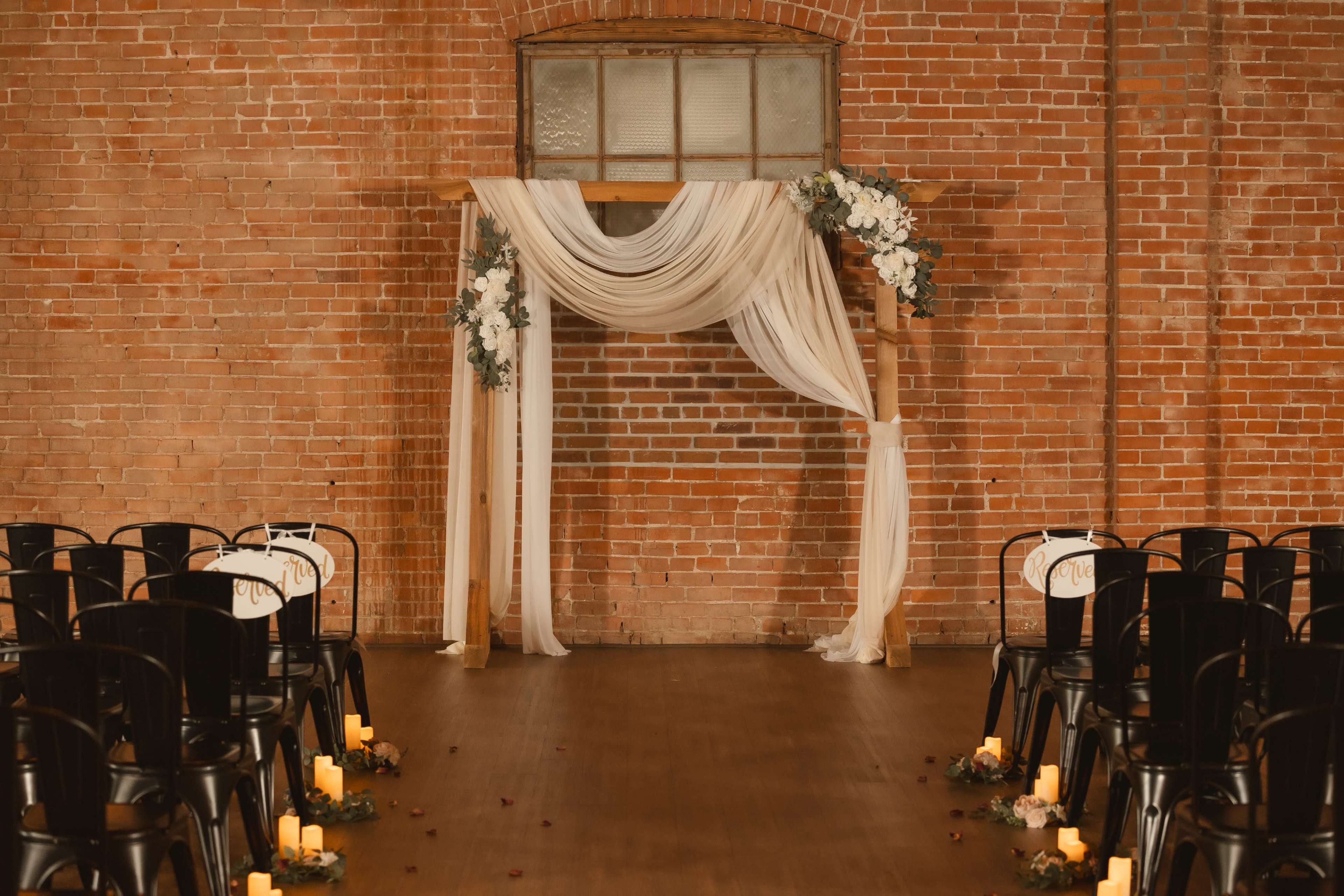A wedding altar adorned with draped fabric and floral arrangements is set against a brick wall, flanked by rows of black chairs and candles on the floor.
