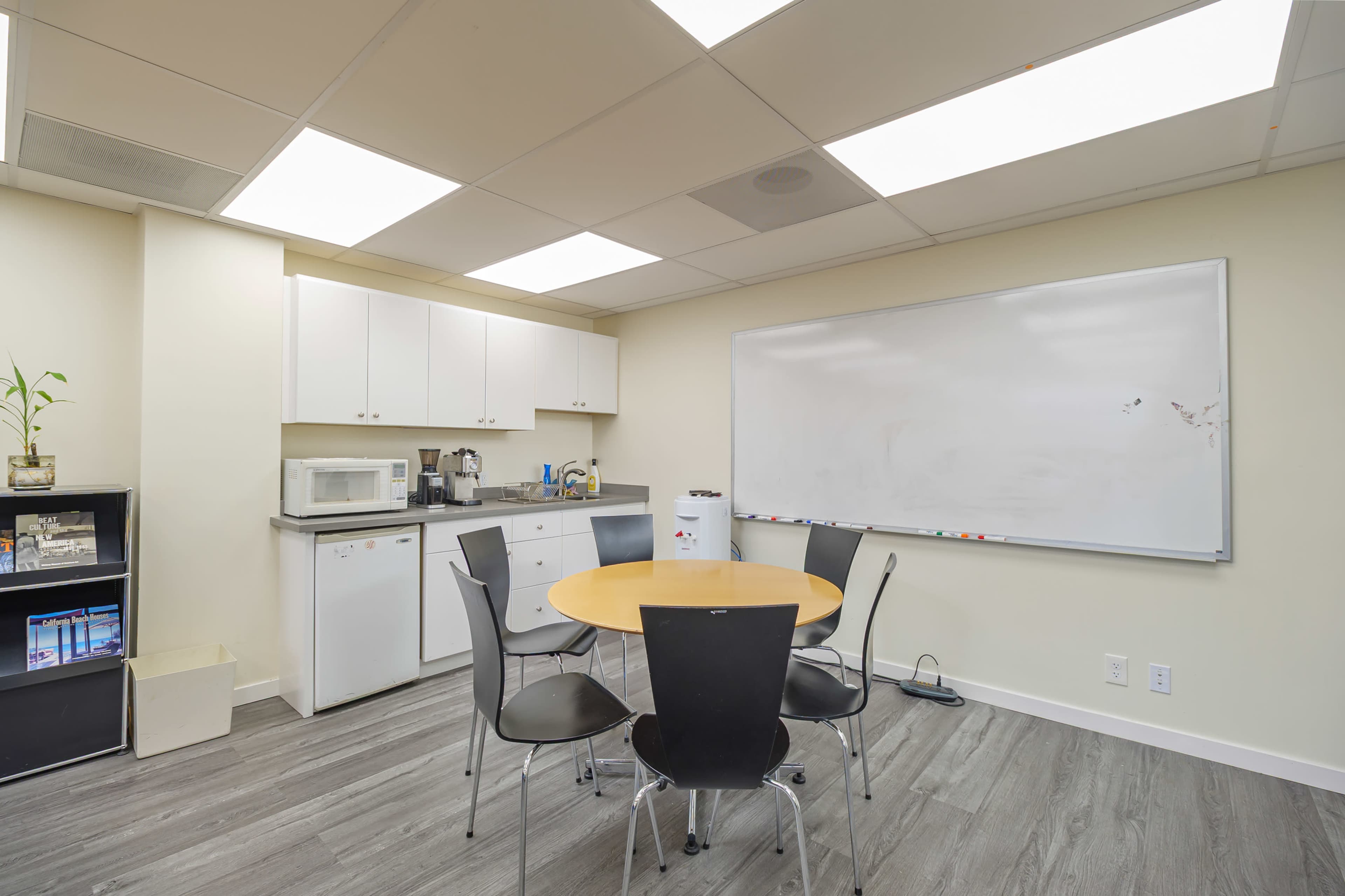 The image shows a small break room or kitchenette area with a round table and four black chairs, a whiteboard on the wall, and kitchen appliances like a microwave and a coffee maker.