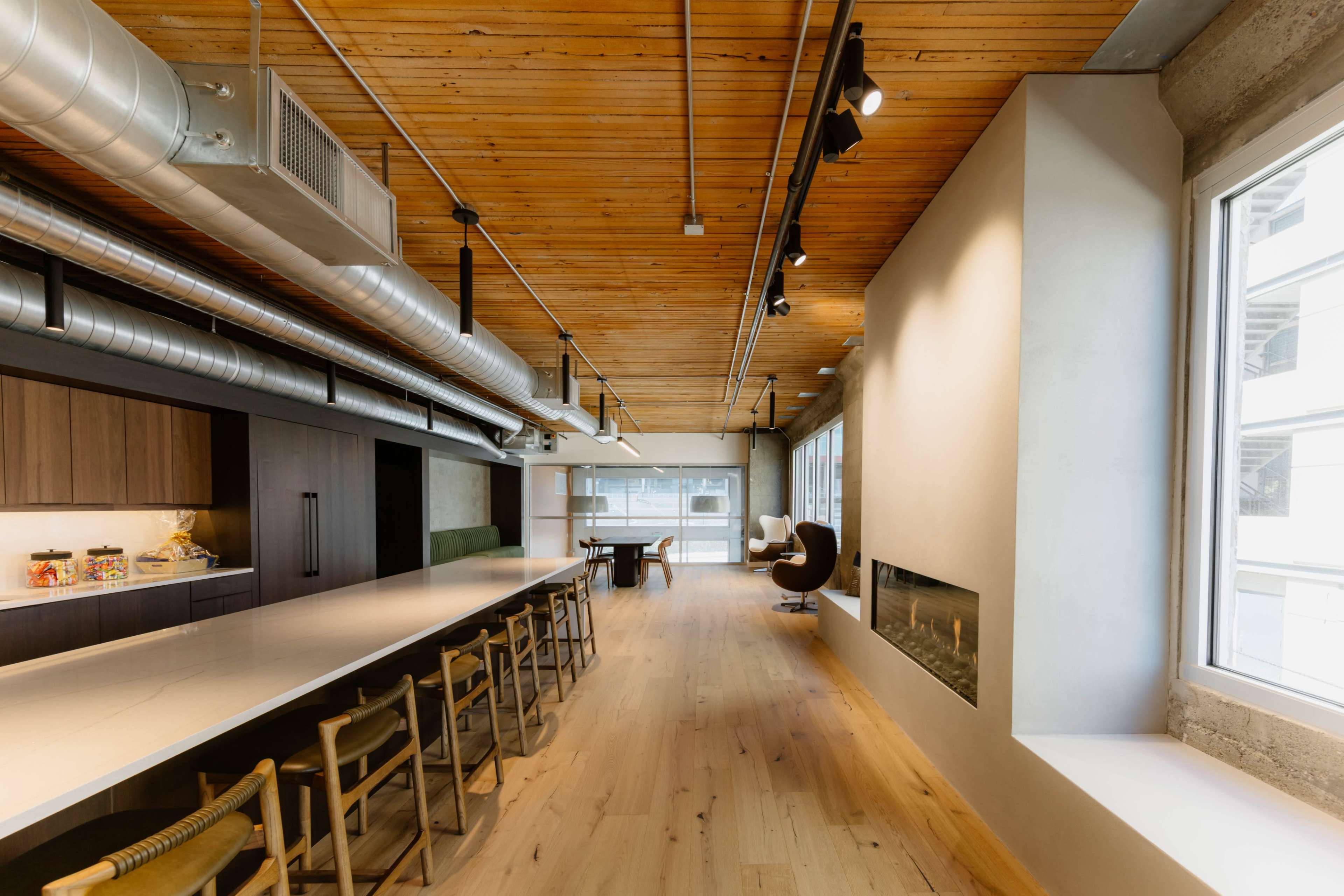 The image shows a modern interior space featuring a long kitchen island, wooden flooring, and large windows, with a dining area and a minimalist fireplace along one wall.