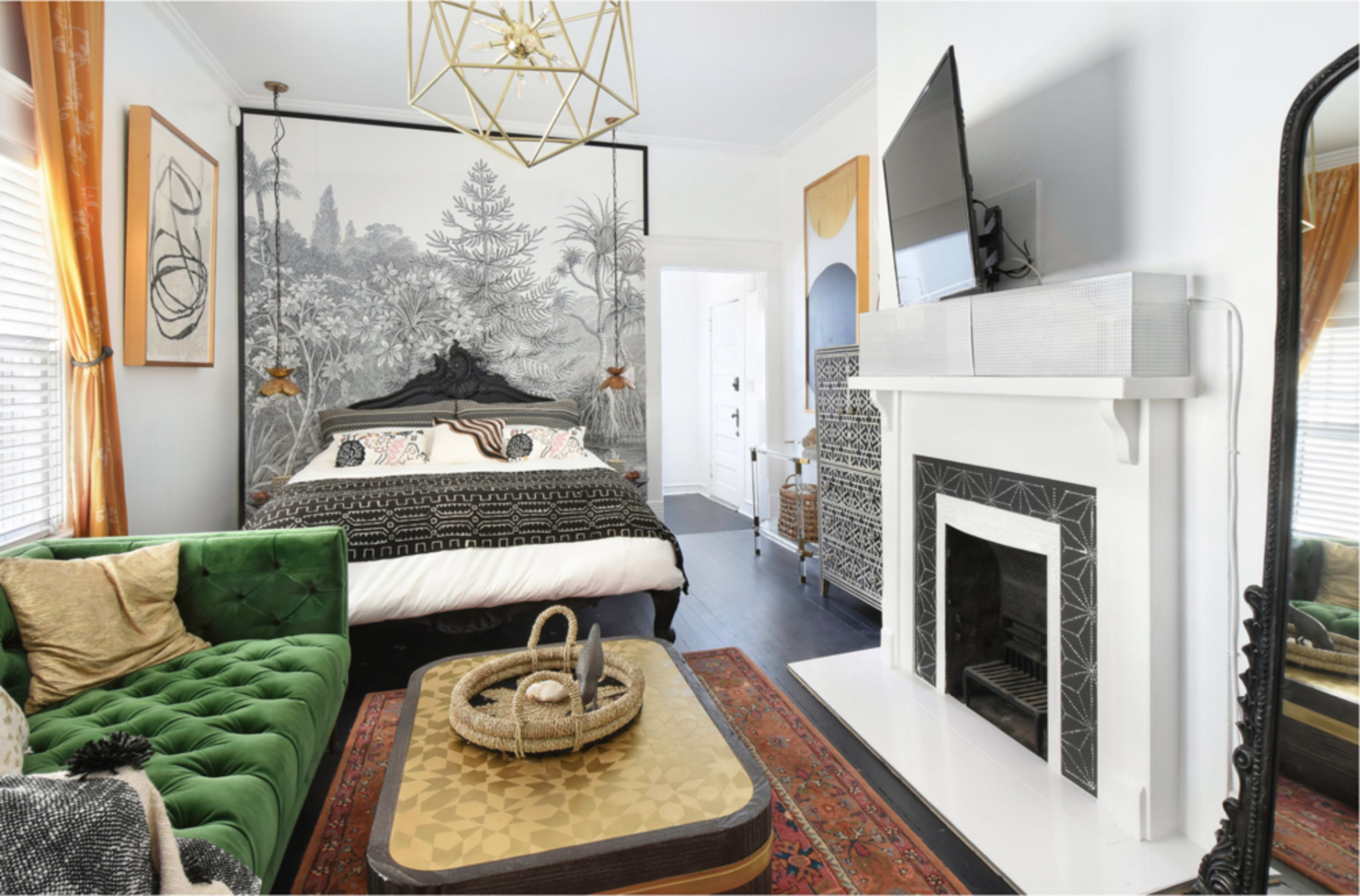 The image depicts a stylish bedroom with a green velvet sofa, a large bed, and a decorative fireplace, complemented by a patterned wall mural and modern lighting fixtures.