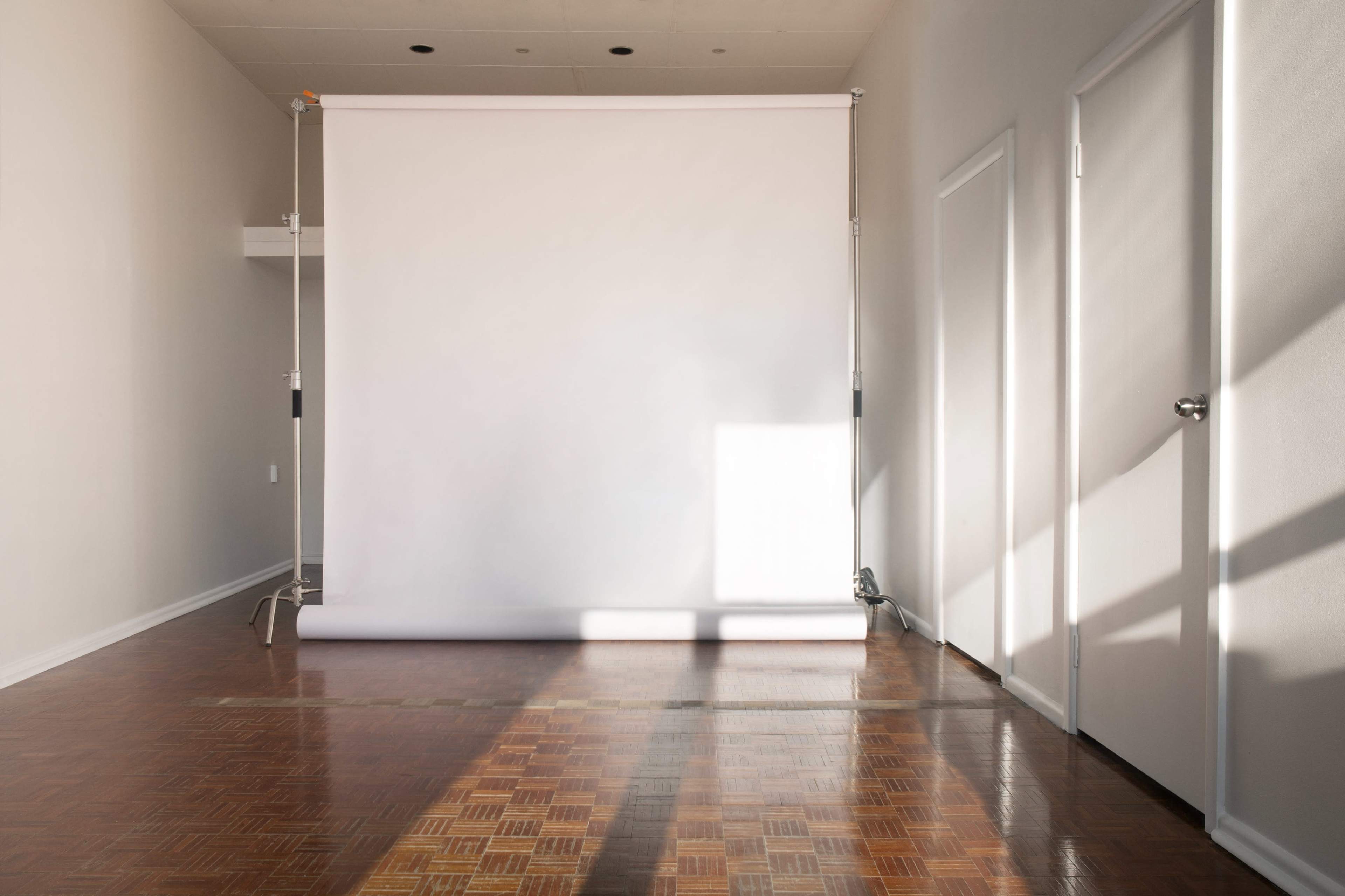 A minimalist studio space with a white backdrop roll, wooden flooring, and two closed doors on adjacent walls.