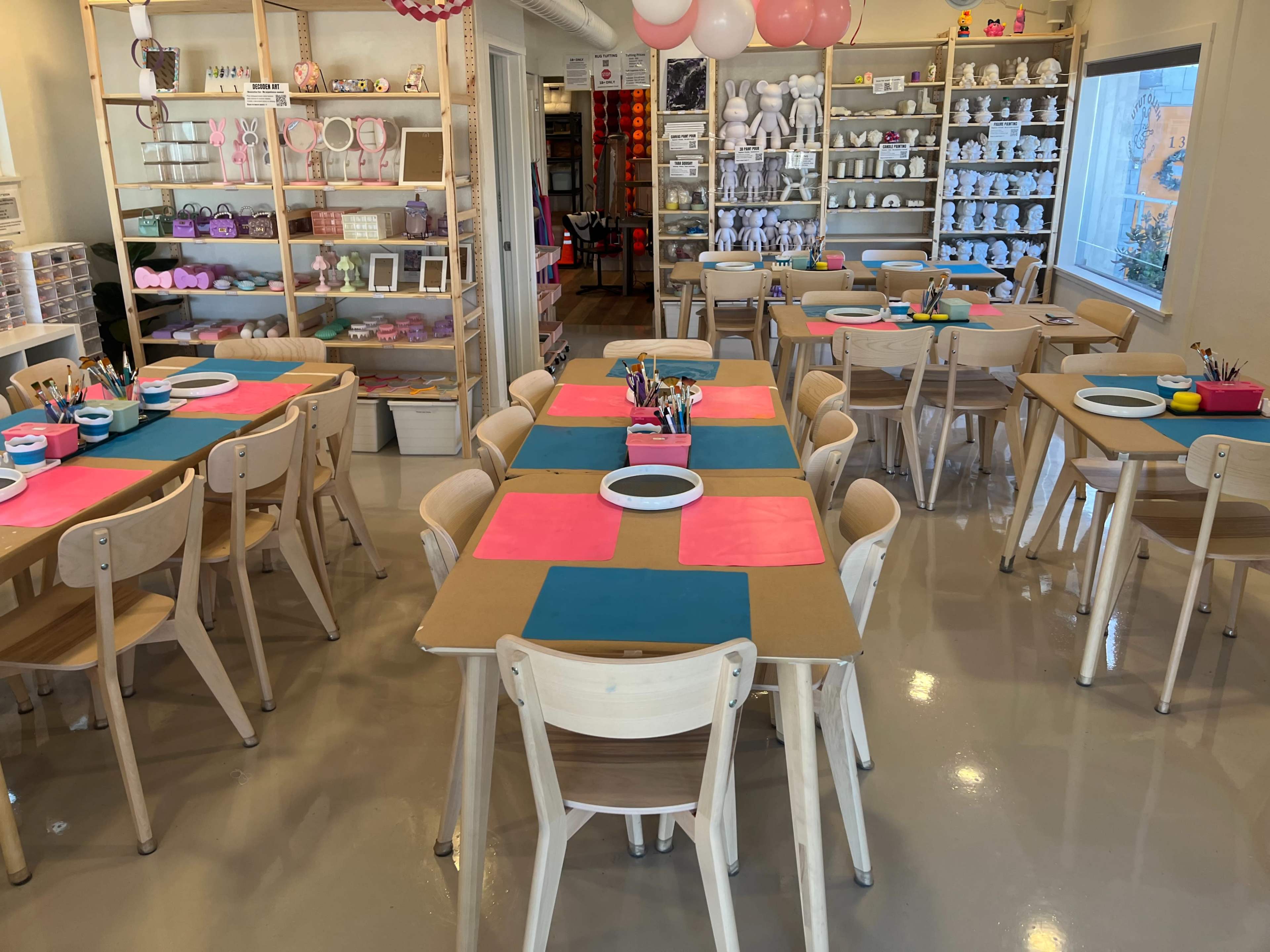 The image shows an art studio with multiple tables set up for painting activities, featuring colored table covers, supplies, and organized shelves displaying various pottery items.