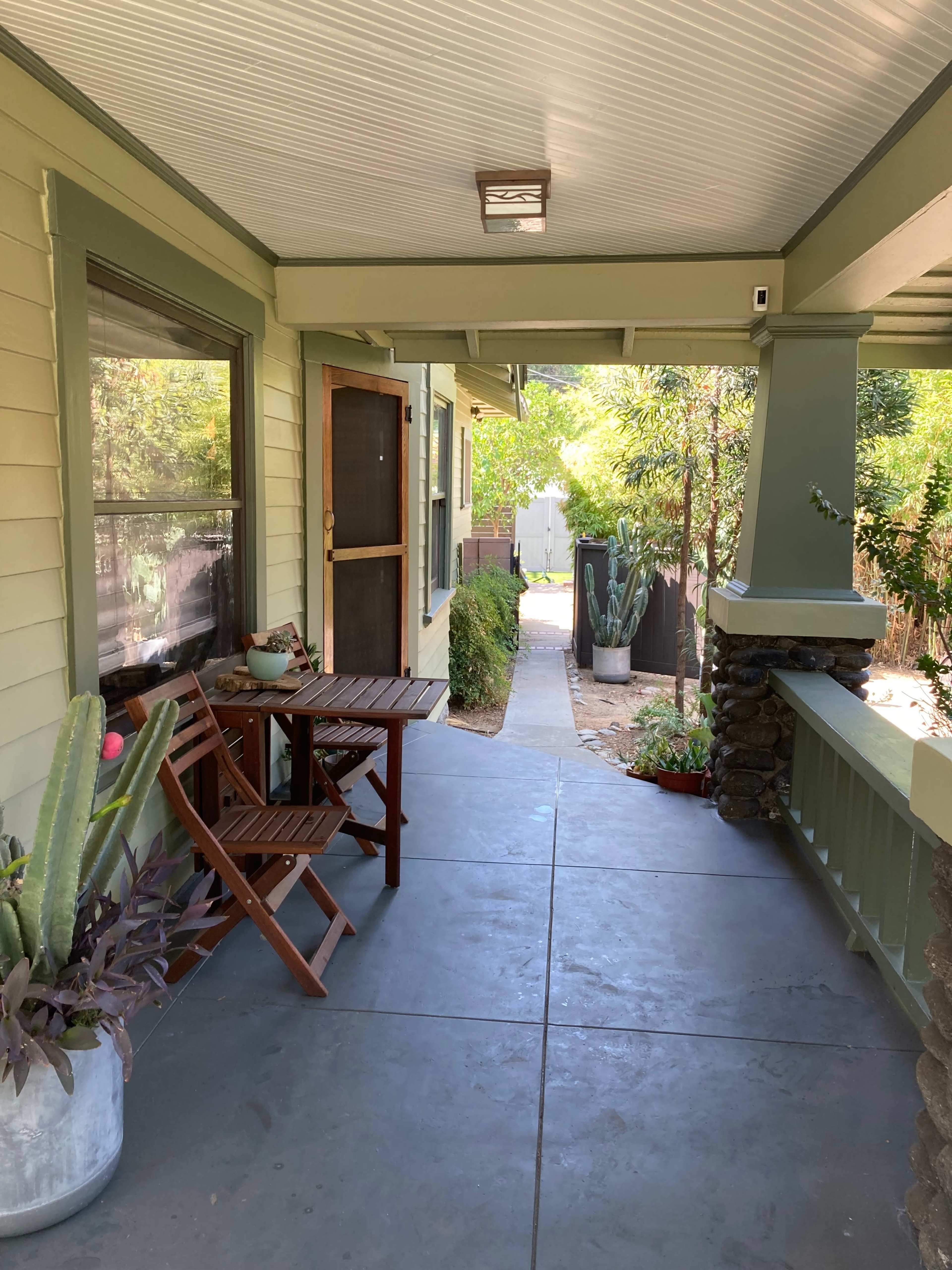 A porch with a wooden folding table and chairs, surrounded by potted plants and a pathway leading to a garden.