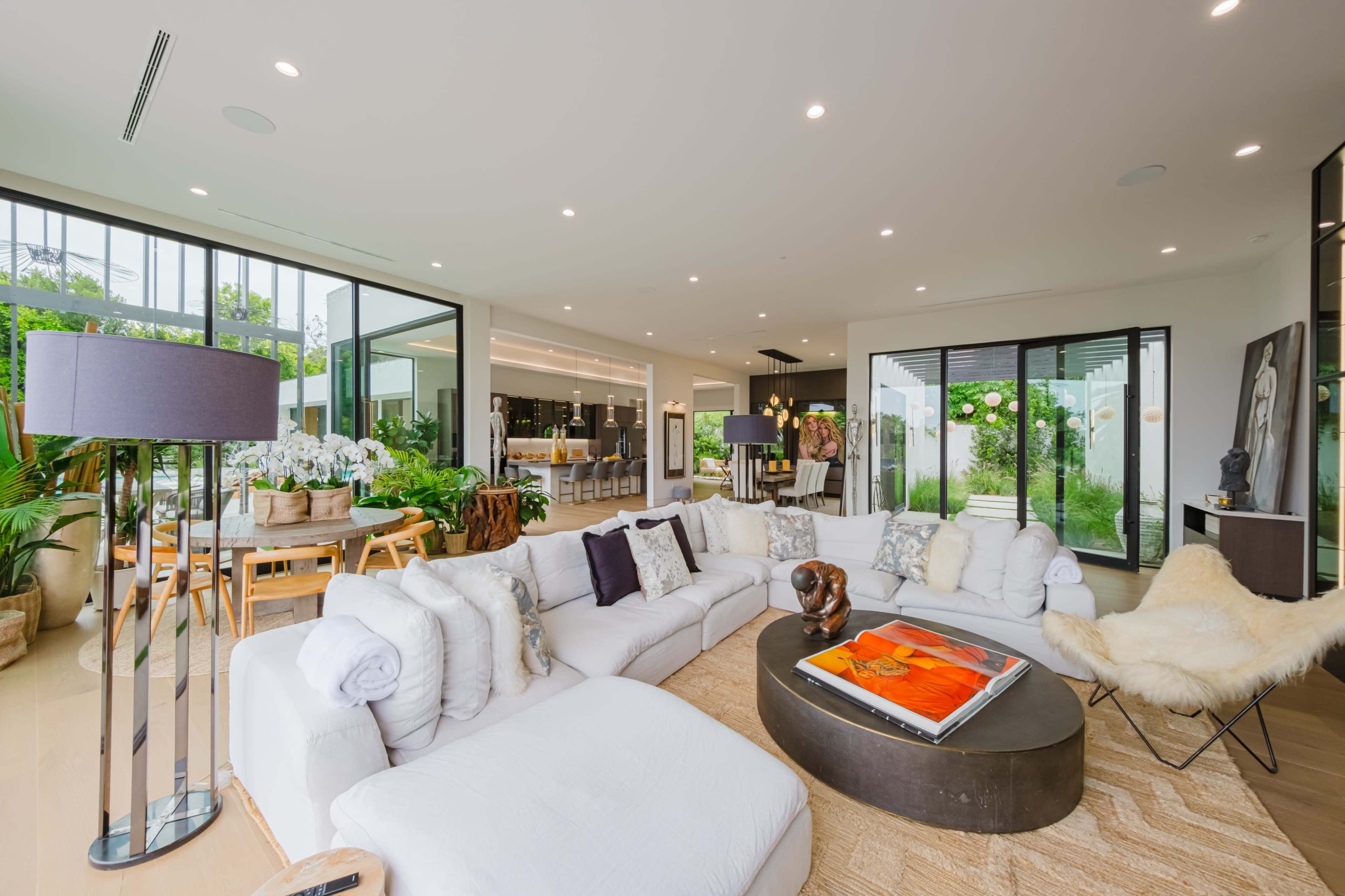 A modern living room with a large white sectional sofa, a round coffee table, and large glass doors leading to an outdoor area filled with greenery.