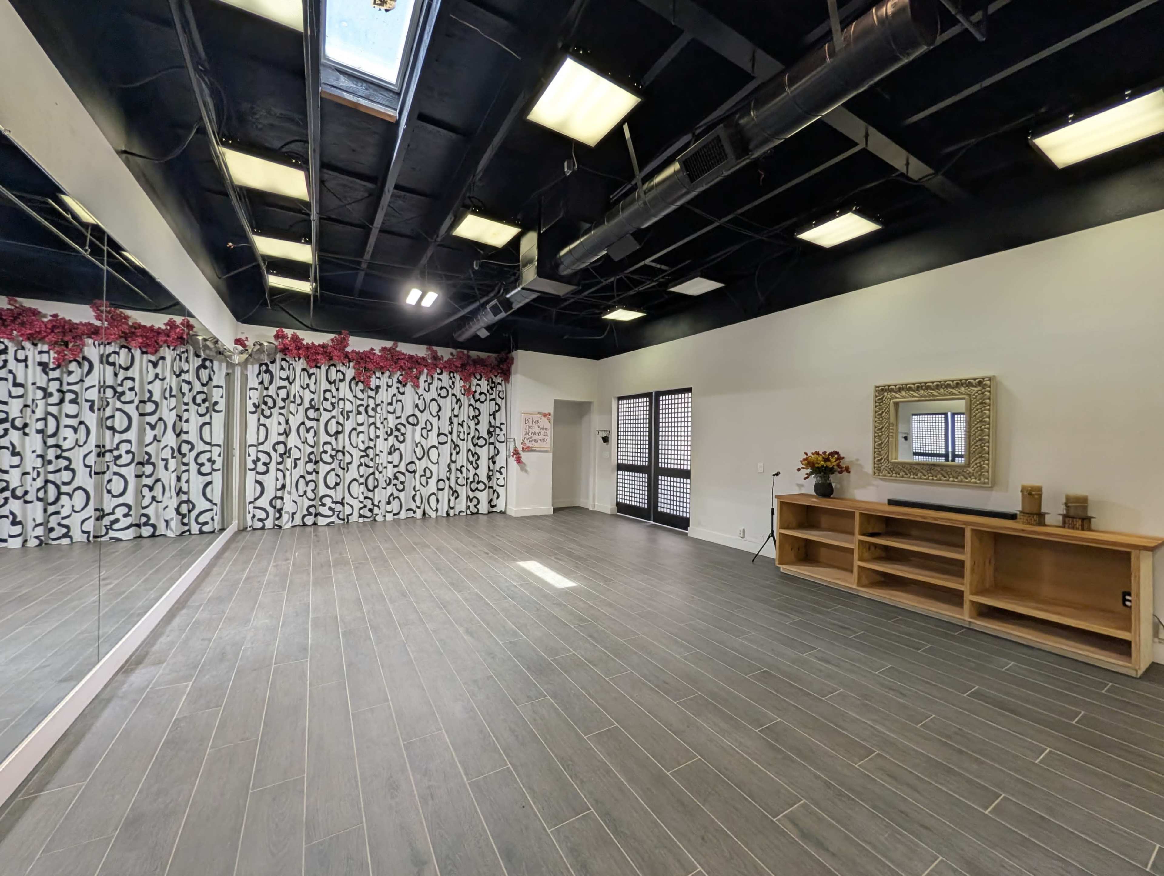 The image shows a spacious, well-lit room featuring a mirrored wall, a wooden storage unit, and floral-accented decor along one side.