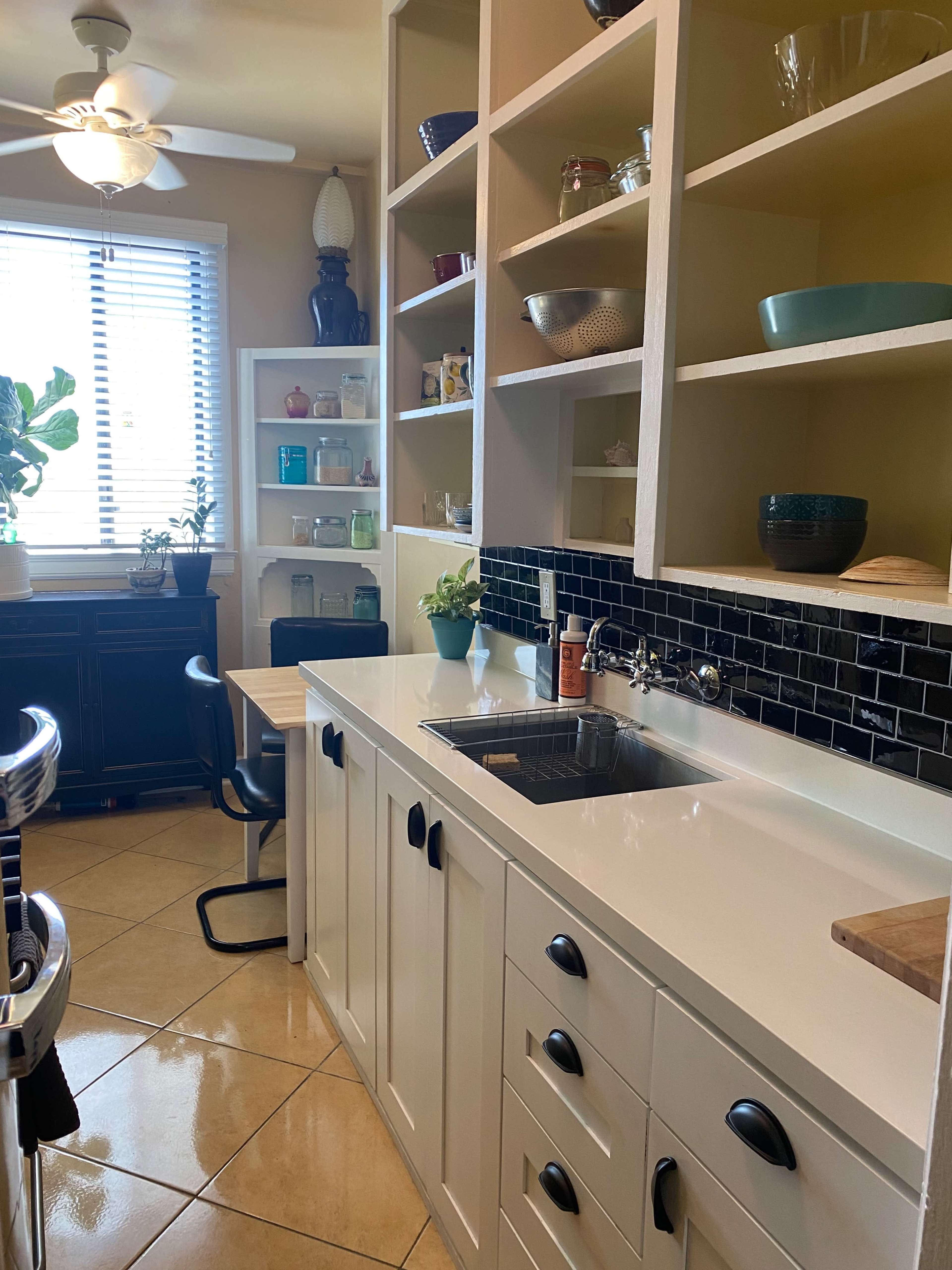 The image shows a clean kitchen area with white cabinetry, a black backsplash, and a small dining table beside a window.