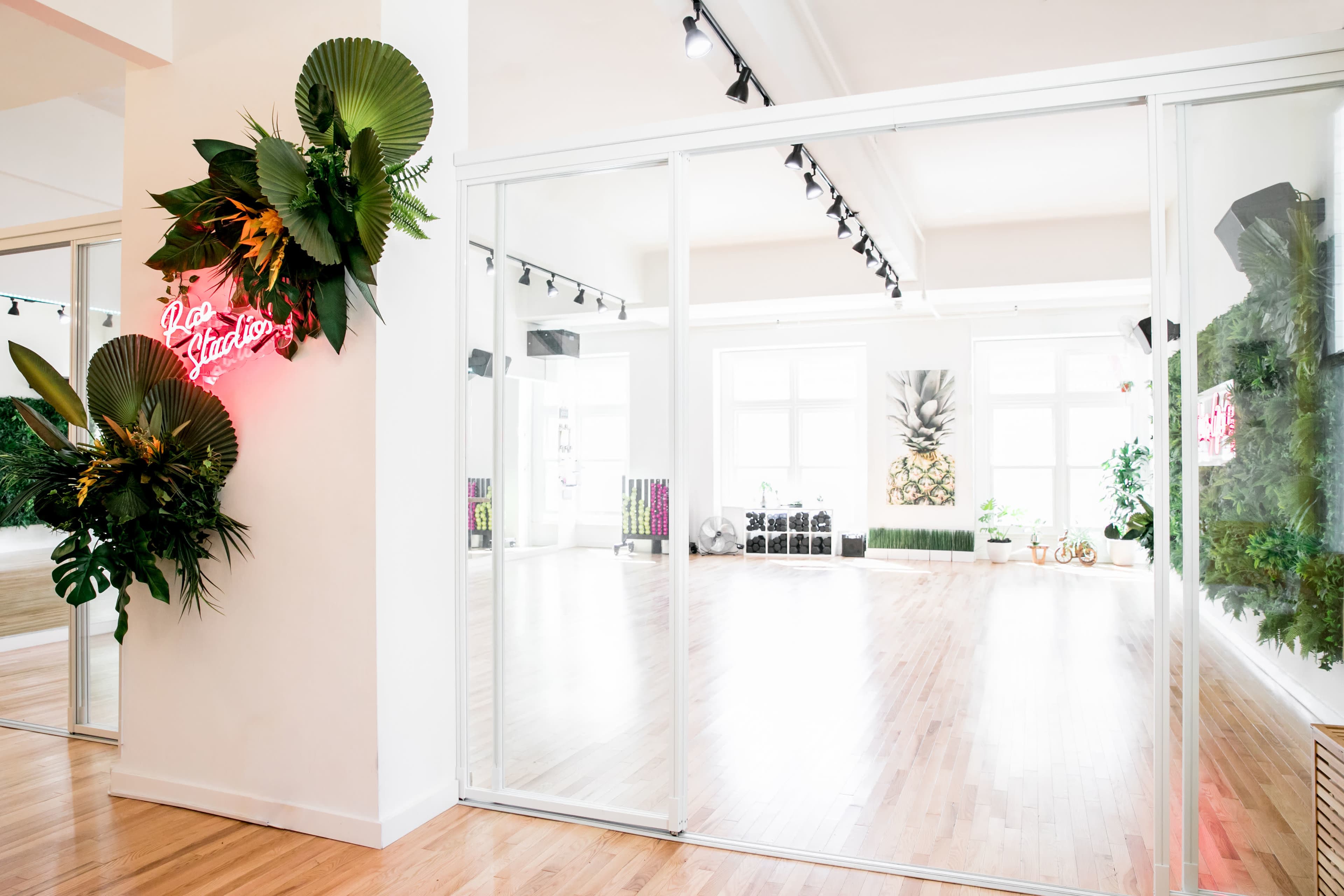 A bright, modern studio space features large glass walls, wooden floors, and greenery decor, with a neon sign on one wall.