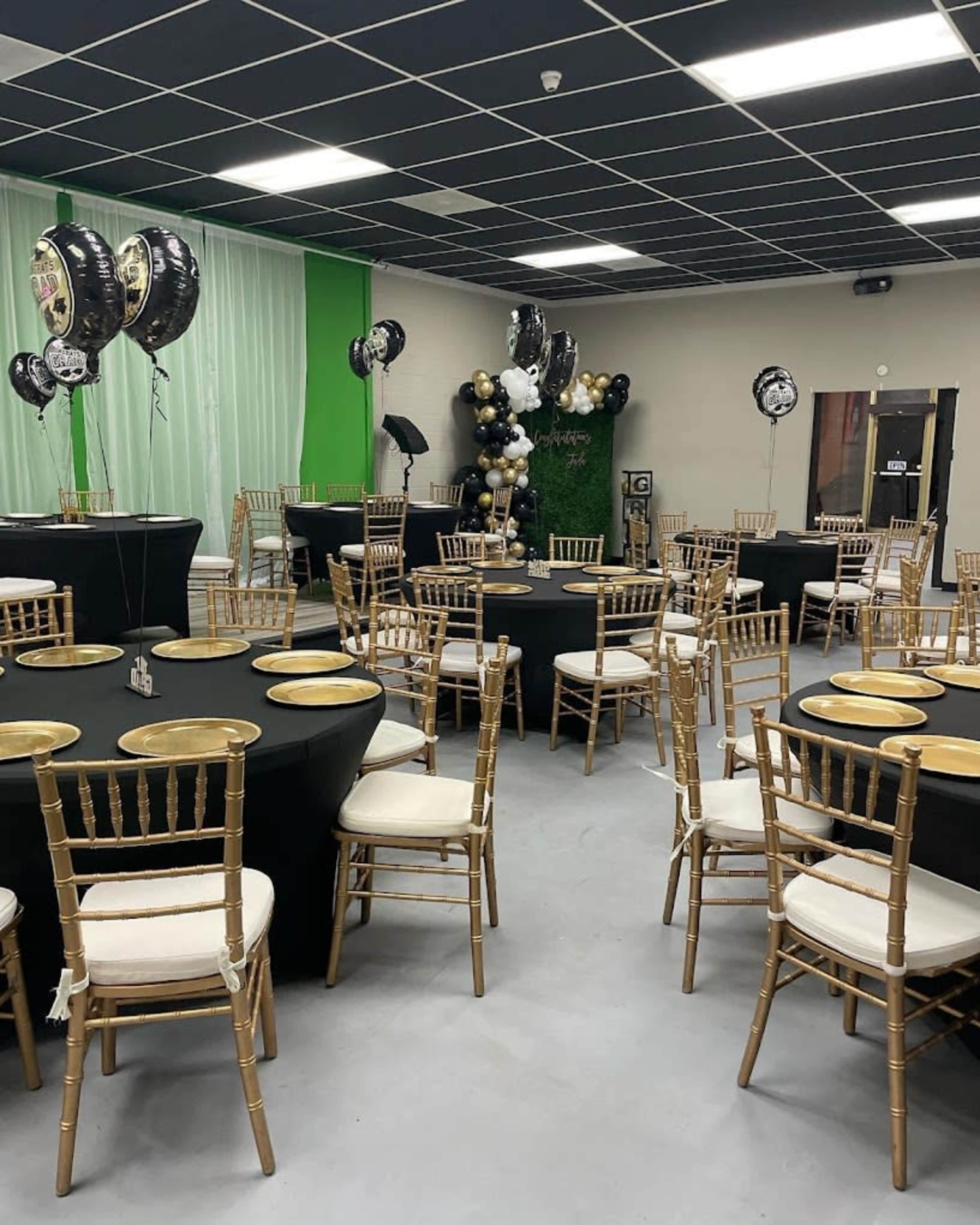The room is set up for an event with black and gold themed tables and decorations, featuring round tables with black tablecloths and gold plates, surrounded by gold chiavari chairs.