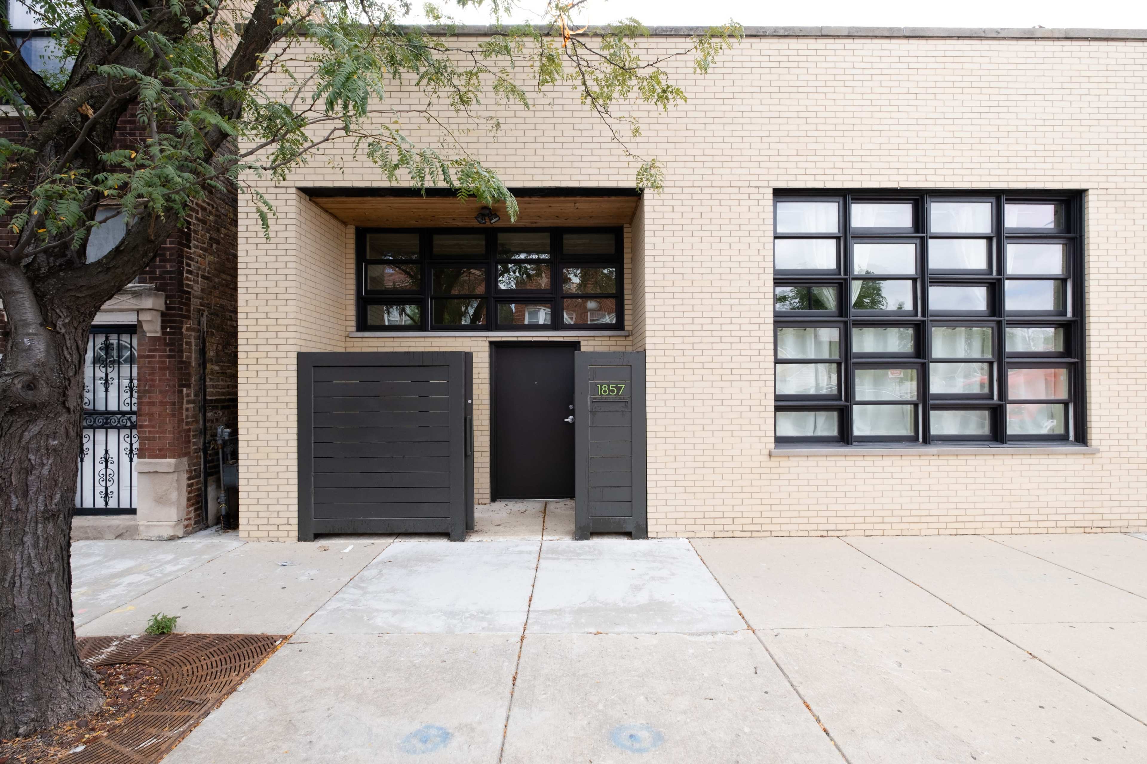 A modern, light-colored brick building with large black-framed windows and a front entrance with a black door, flanked by two dark wooden storage units.