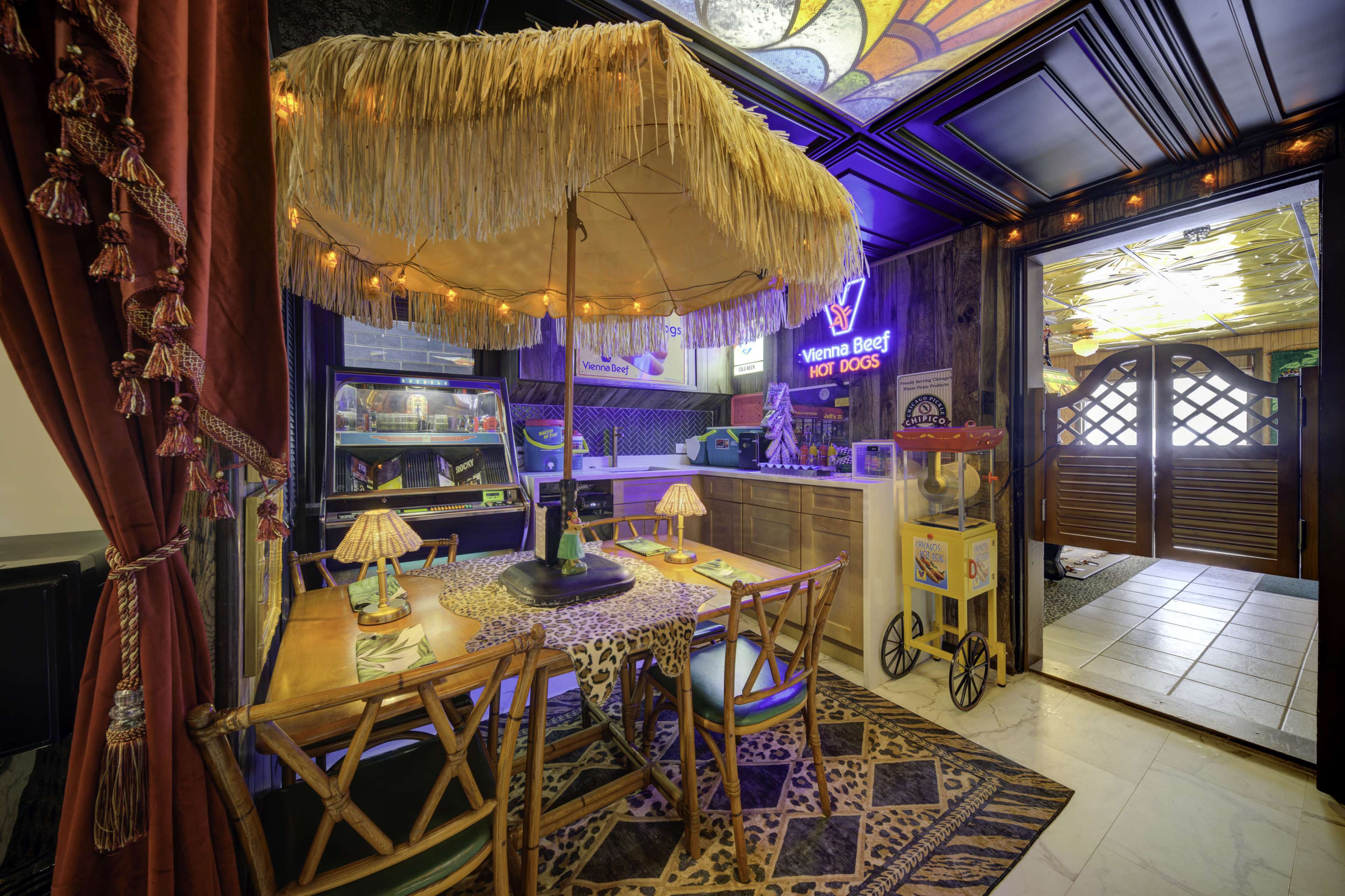The image shows a tropical-themed dining area featuring a table under a thatched umbrella, with colorful decor and an entrance partially visible in the background.
