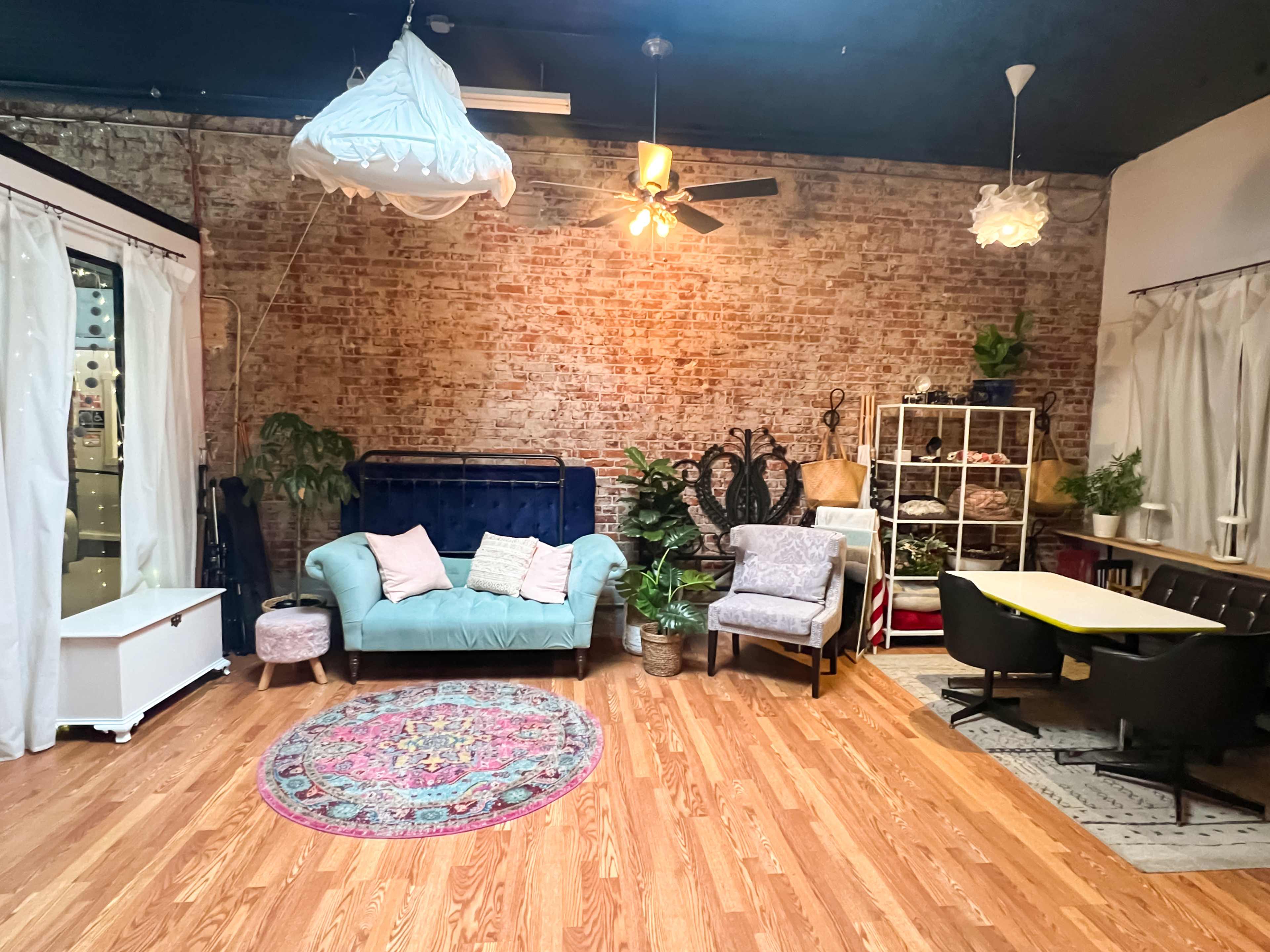 A spacious room with exposed brick walls, featuring a mix of seating arrangements, houseplants, and decorative rugs.