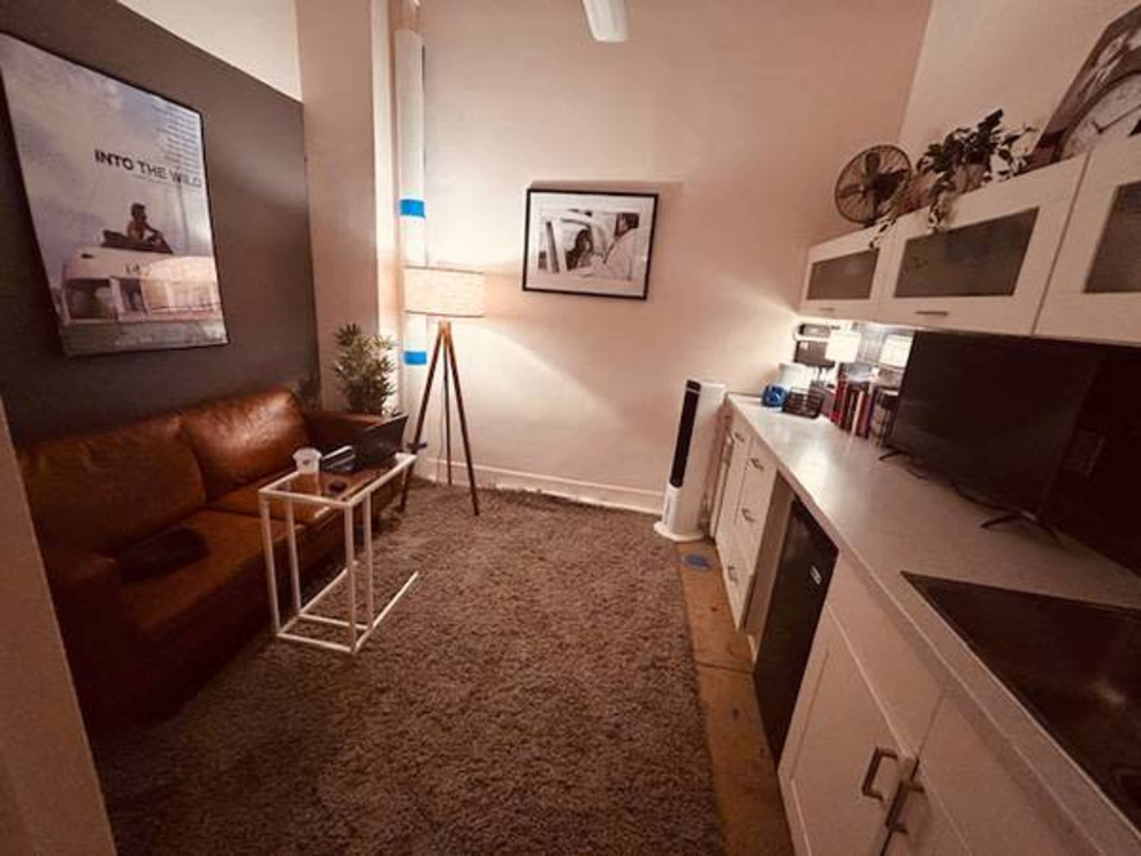 The image shows a small, cozy room featuring a brown leather sofa, a standing lamp, and a minimalist kitchen area with white cabinets and a countertop.
