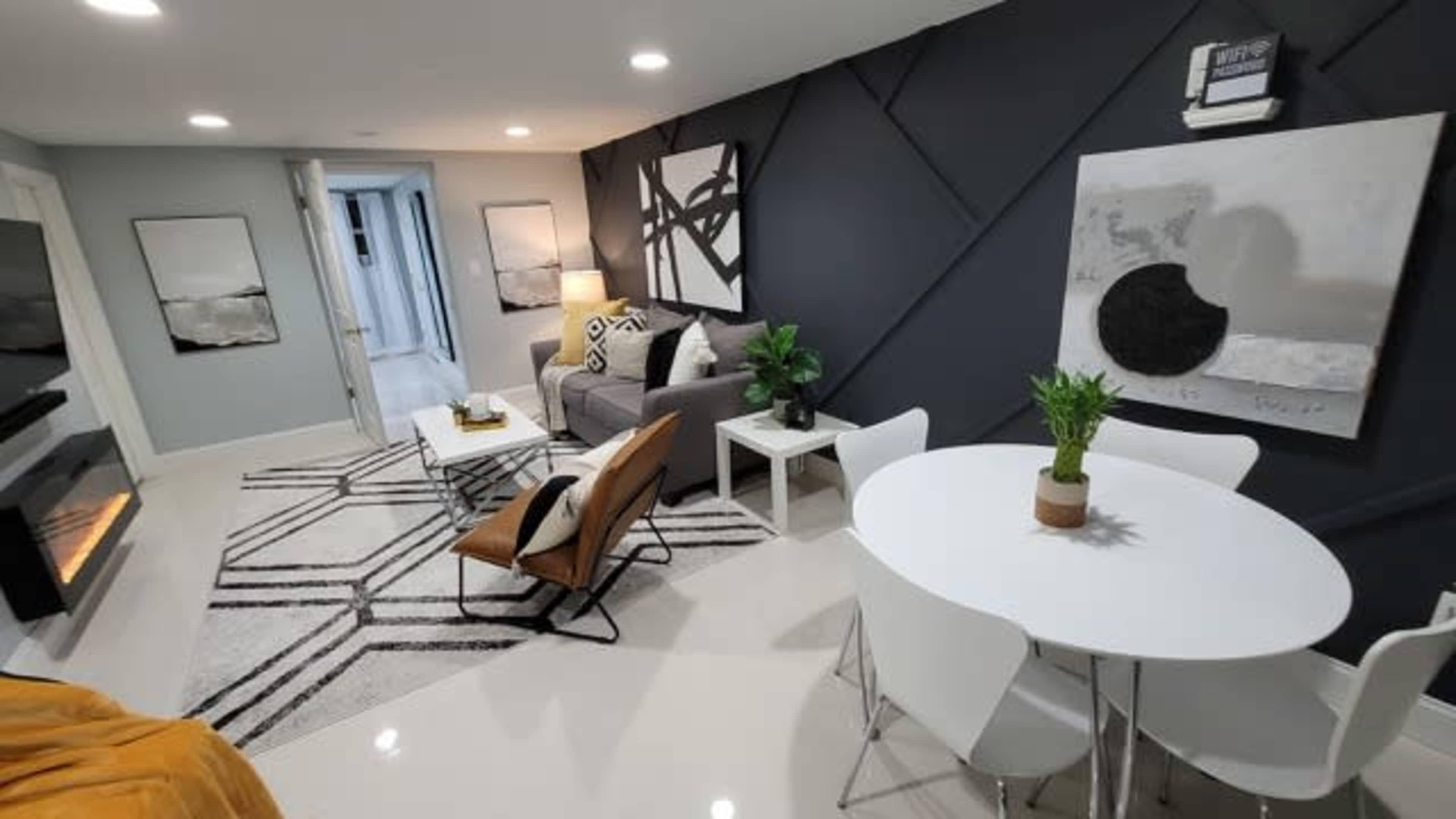 A modern living room features a grey sofa, a minimalist coffee table, a white dining table with chairs, and abstract wall art against a dark accent wall.