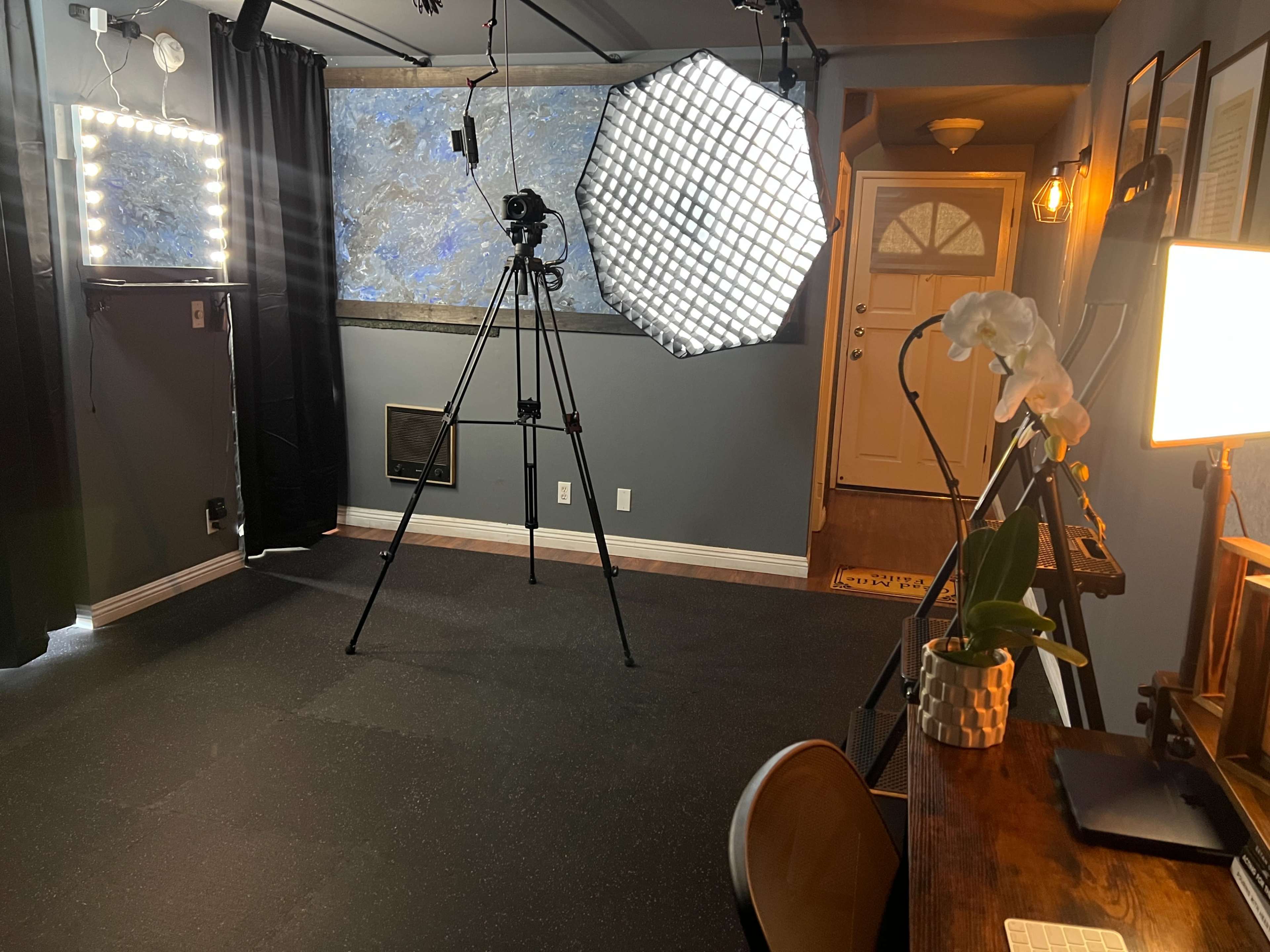 A well-lit room featuring a camera on a tripod, softbox lighting, a desk with a lamp and plants, and a door in the background.