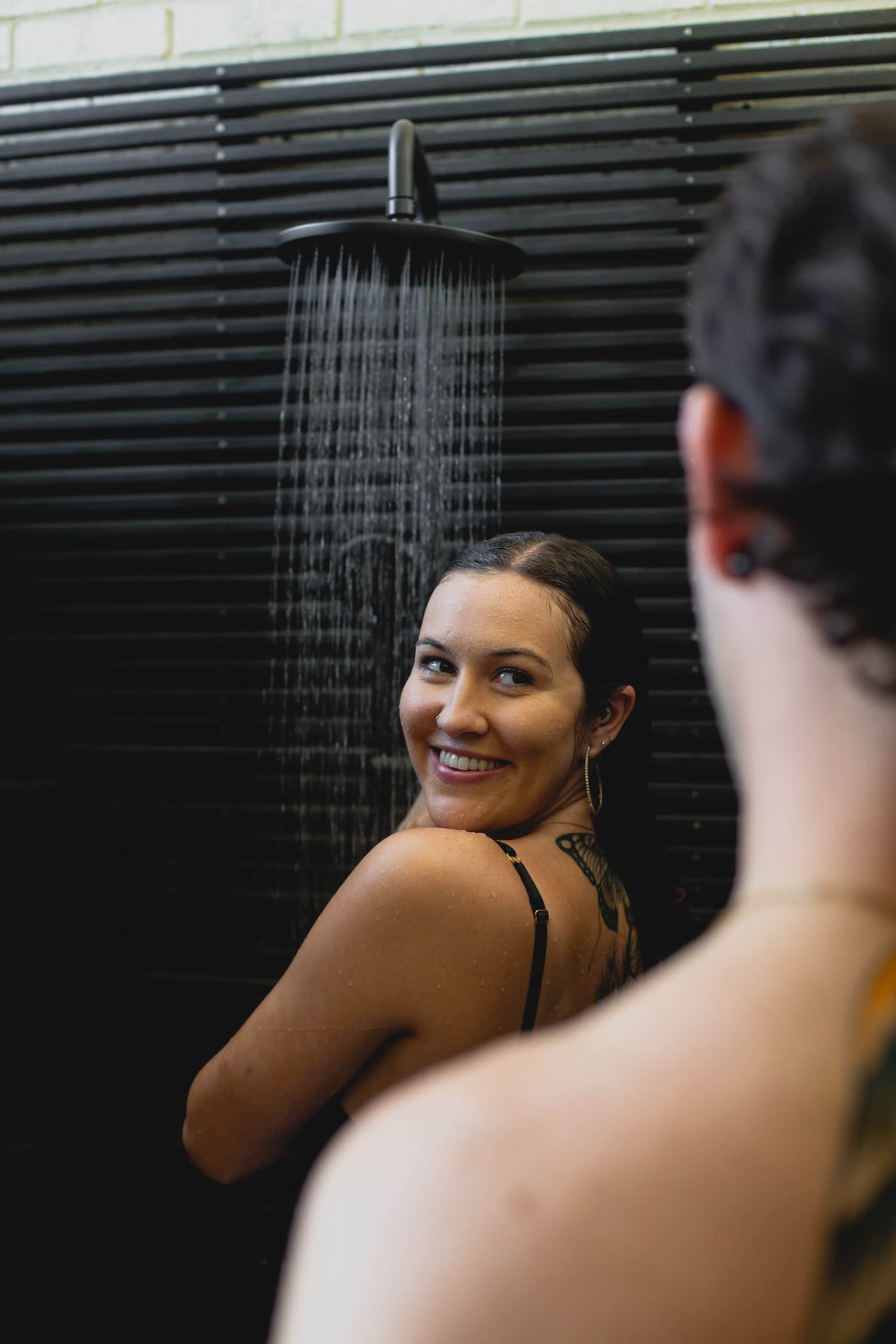 A woman with wet hair smiles while standing under a shower, as a person partially visible in the foreground looks on.