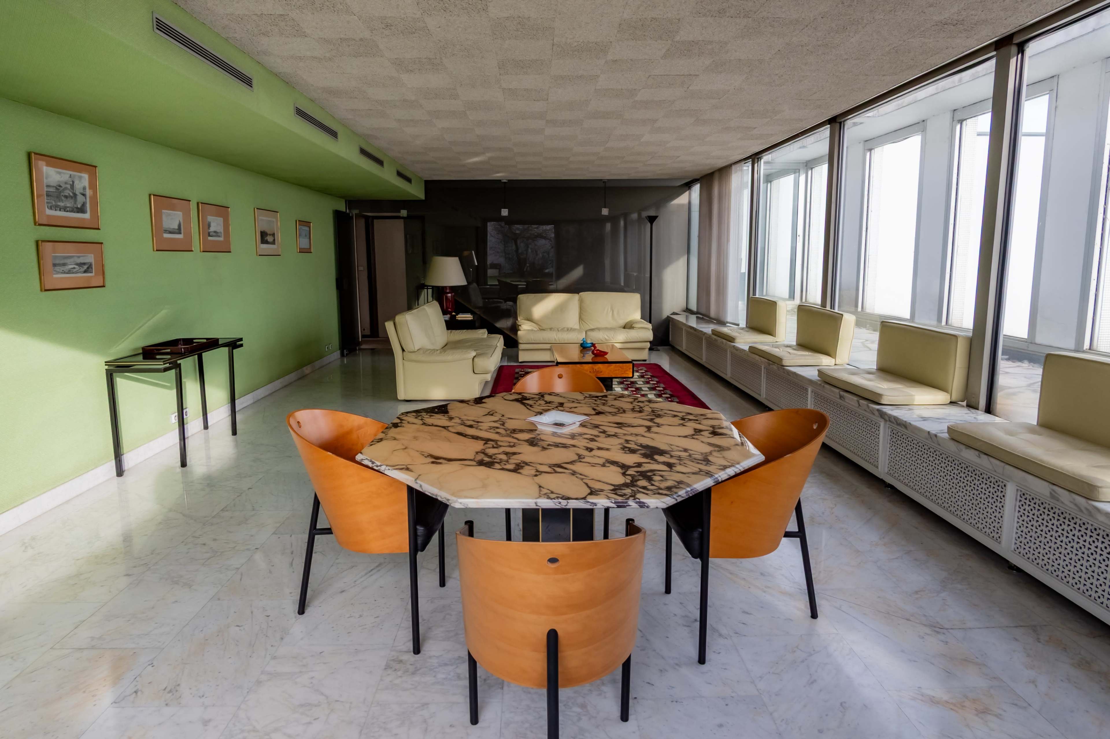 A modern living space features a marble-topped table surrounded by four wooden chairs, with a separator of large windows and light-colored furniture in the background.