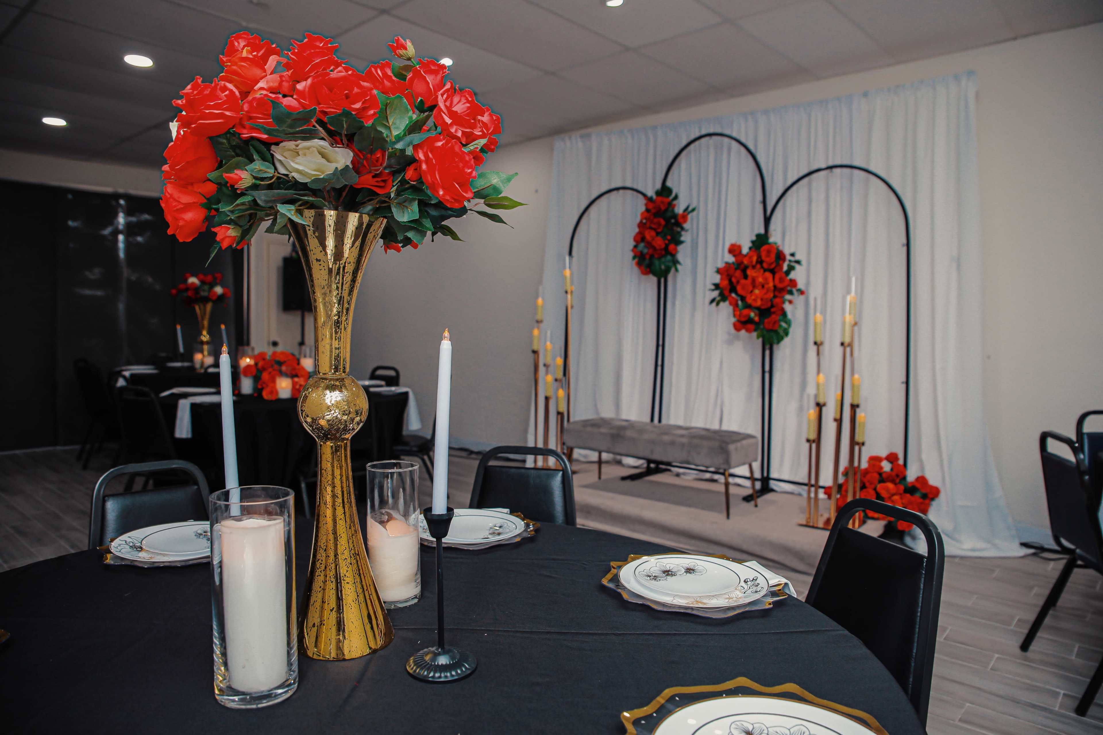 A decorated event space features tables set with black tablecloths, floral centerpieces, and a backdrop adorned with red flowers and candles.