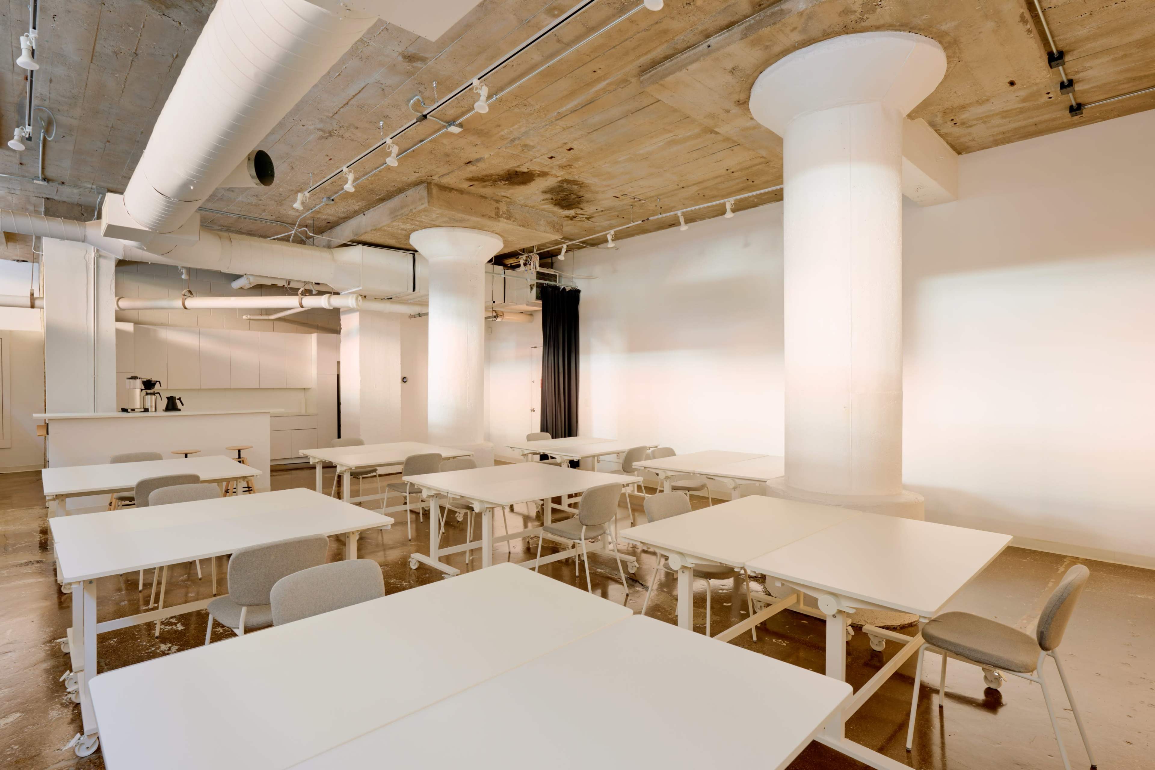 The image shows a spacious, minimalist classroom with white tables arranged in a grid and large concrete columns supporting the ceiling.