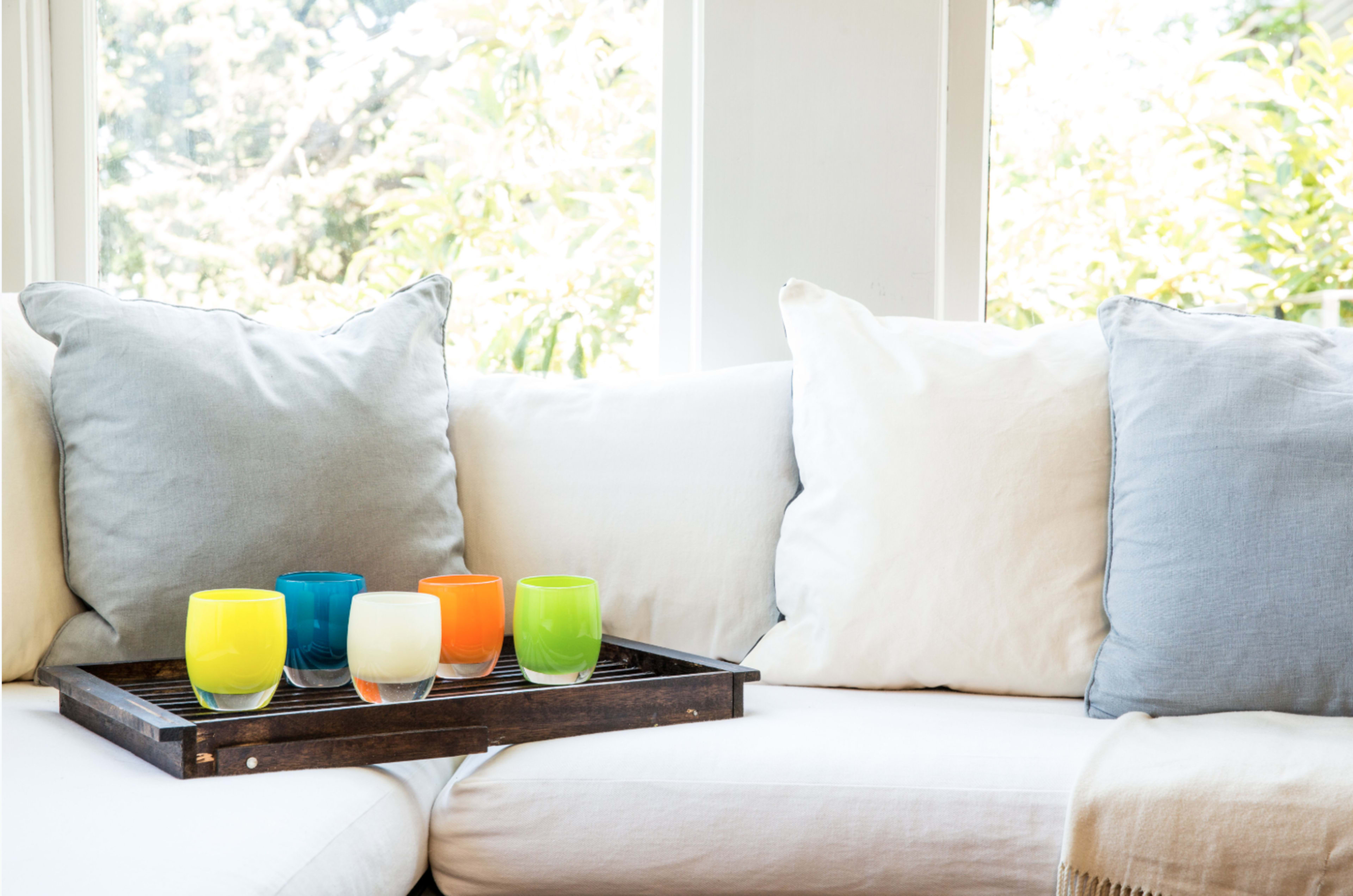 A wooden tray holds five colorful glasses on a light-colored couch adorned with gray and white pillows, set against a backdrop of large windows.