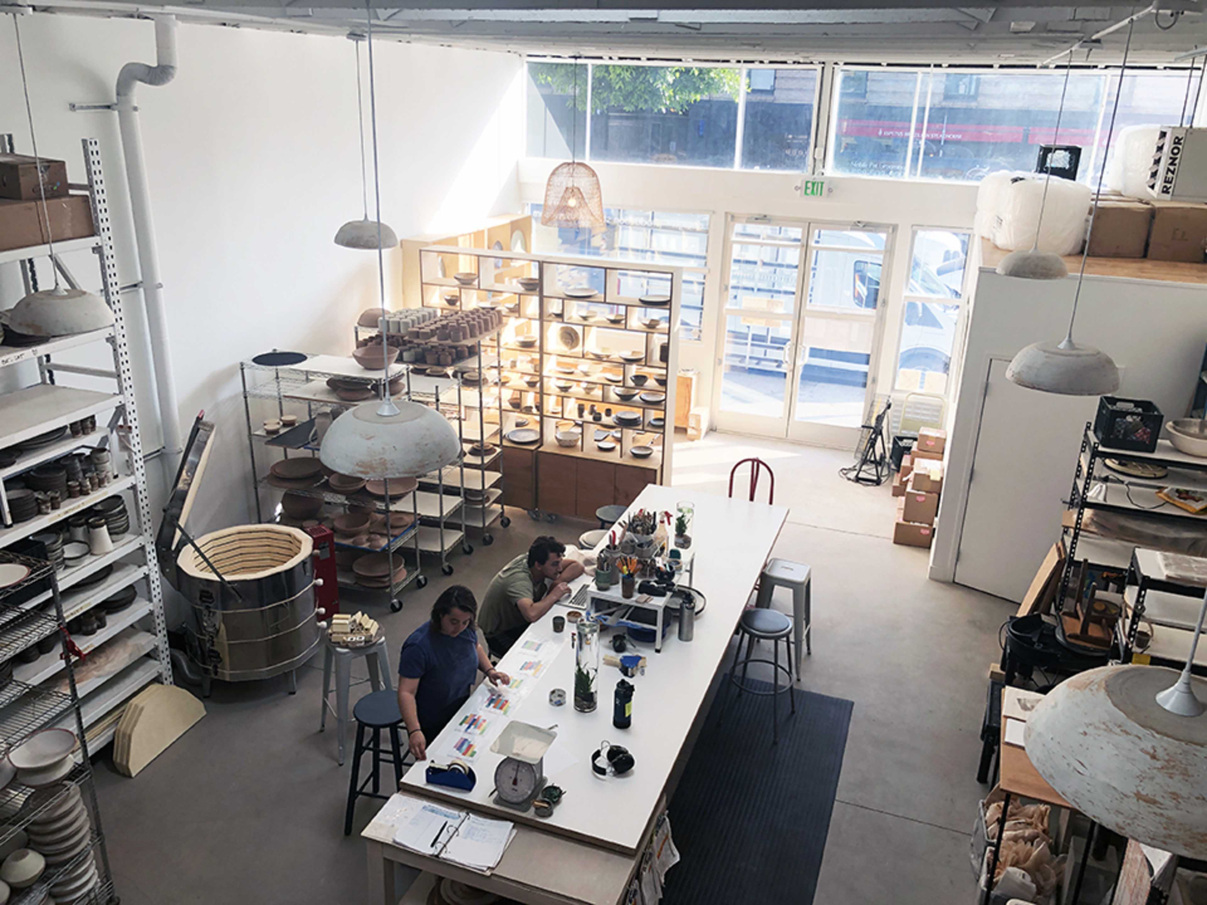 A pottery studio with two people working at a central table surrounded by shelves filled with ceramic pieces and tools.