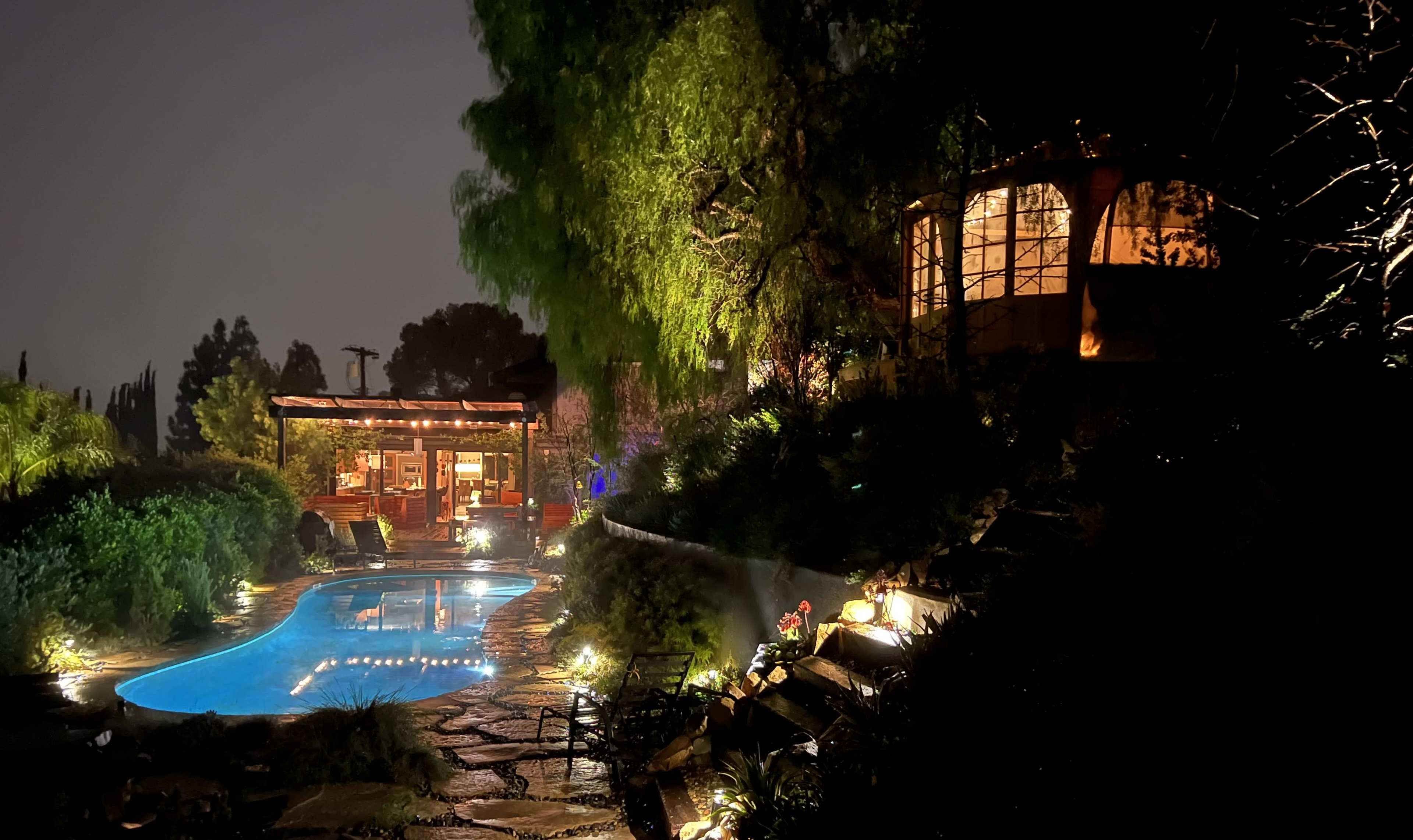 The scene depicts a illuminated backyard pool area at night, surrounded by greenery and featuring a charming wooden structure in the background.