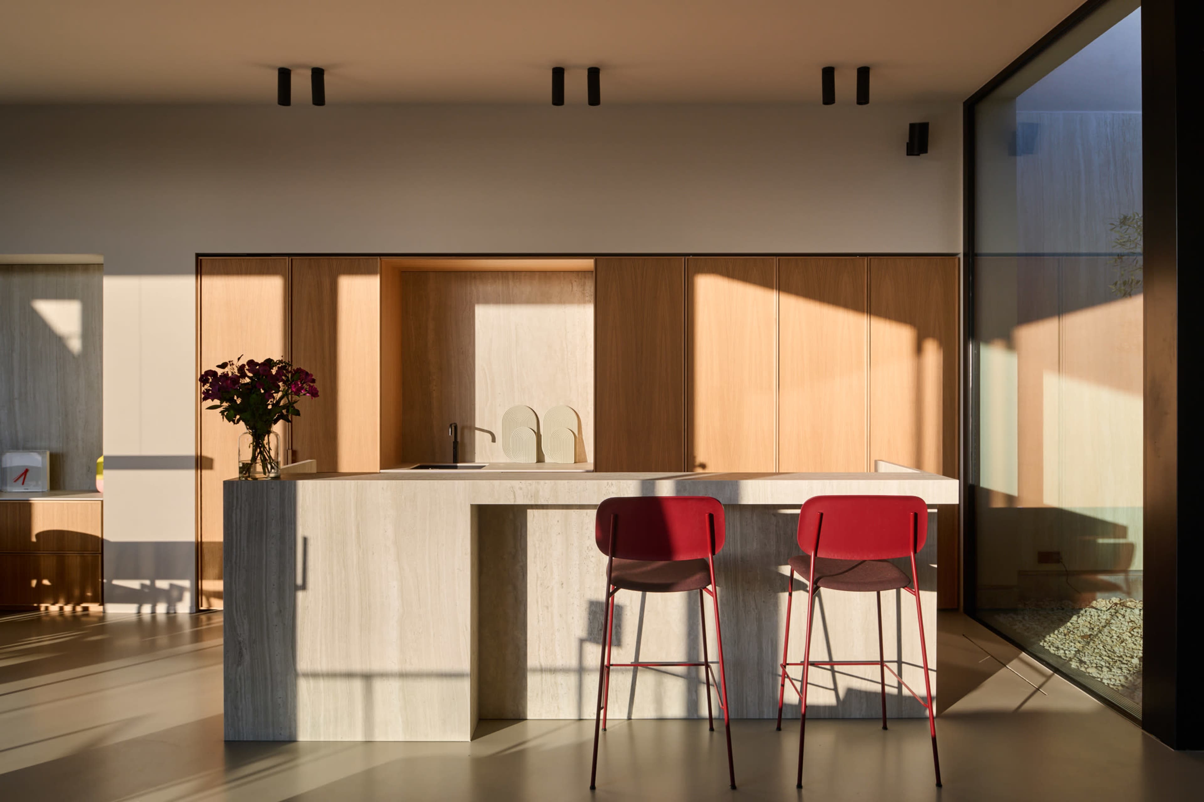 A modern kitchen features a stone island with two red bar stools and wooden cabinets illuminated by natural light.