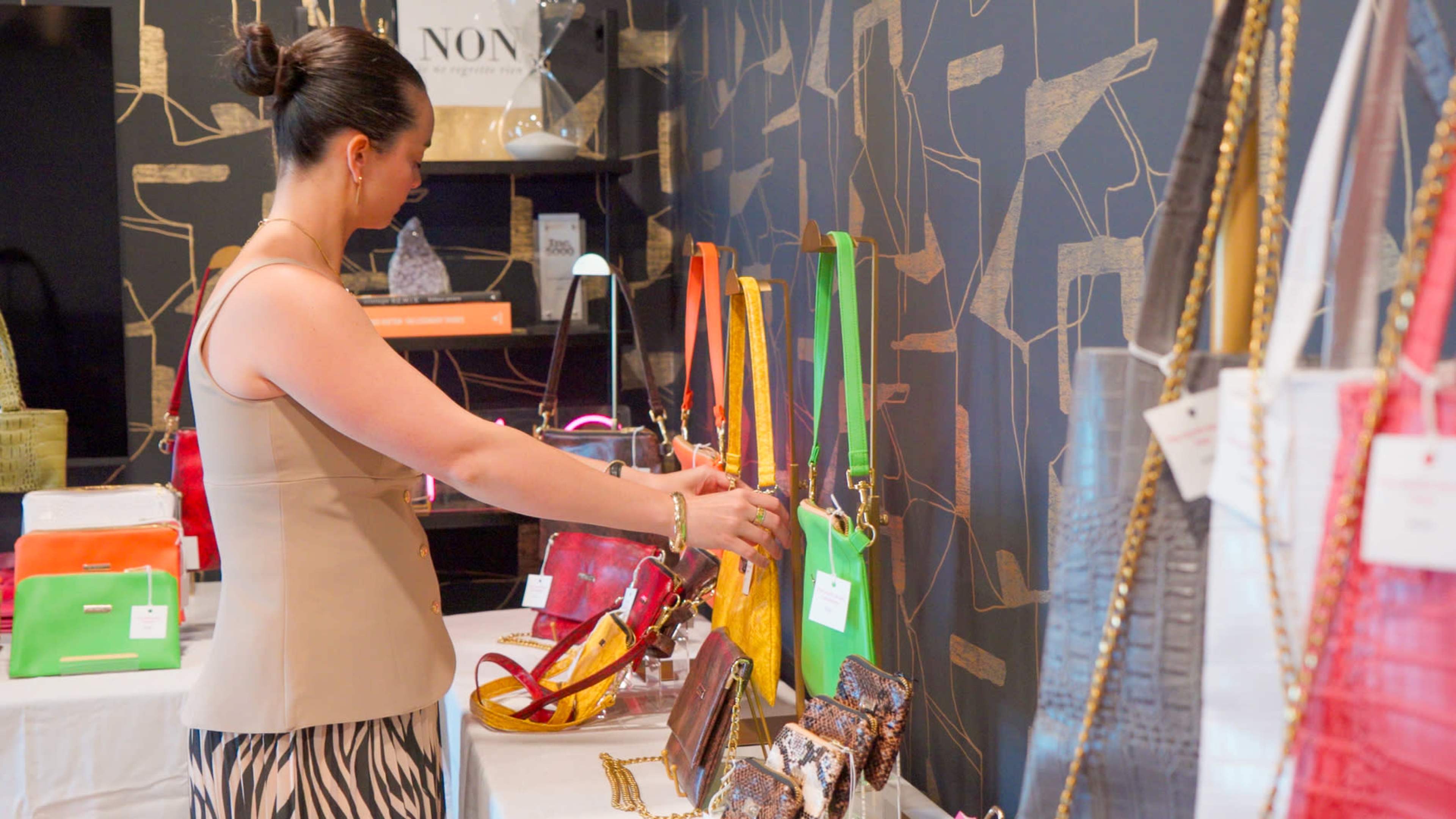 A woman inspects colorful handbags displayed on a table in a retail store.